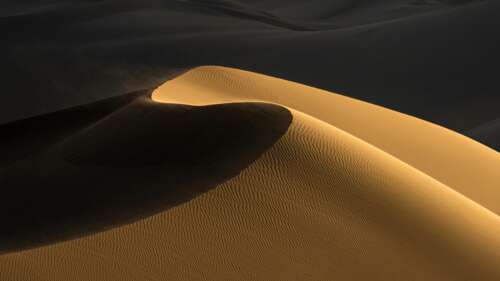 A panoramic landscape of desert dunes during golden hour, featuring sunlight hitting... wallpaper