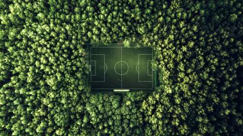 An aerial drone view of a symmetrical soccer field surrounded by a dense, lush green... wallpaper