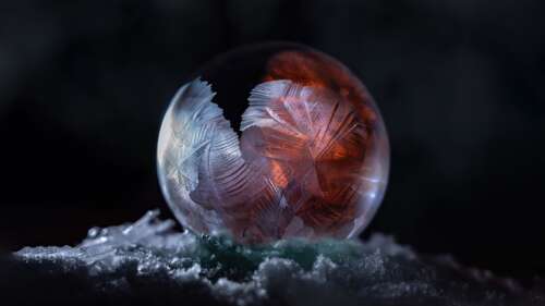 A close-up macro photograph of an iridescent soap bubble resting among white snowflakes... wallpaper
