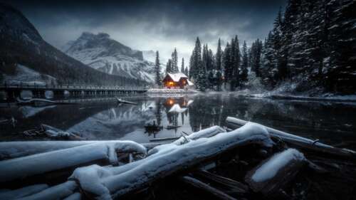 A cozy wooden cabin surrounded by snowy trees and a frozen lake with snow-capped... wallpaper