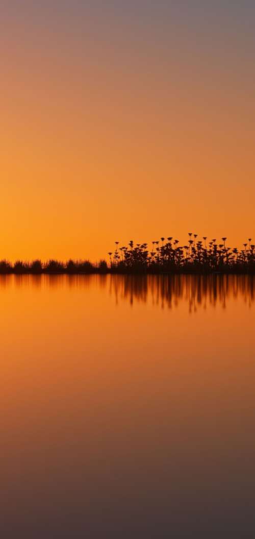 A surreal landscape featuring a vibrant orange sunset sky and silhouetted land reflected... wallpaper