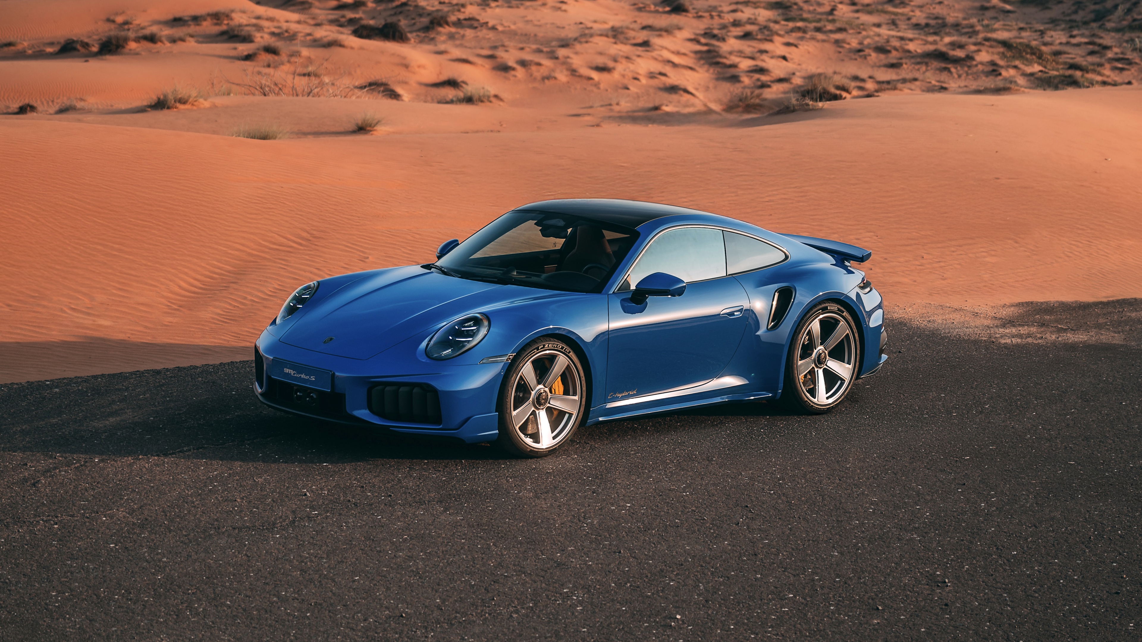 A sleek 2026 Porsche 911 Turbo S parked on the sand dunes of a vast desert under bright, cinematic sunlight.
