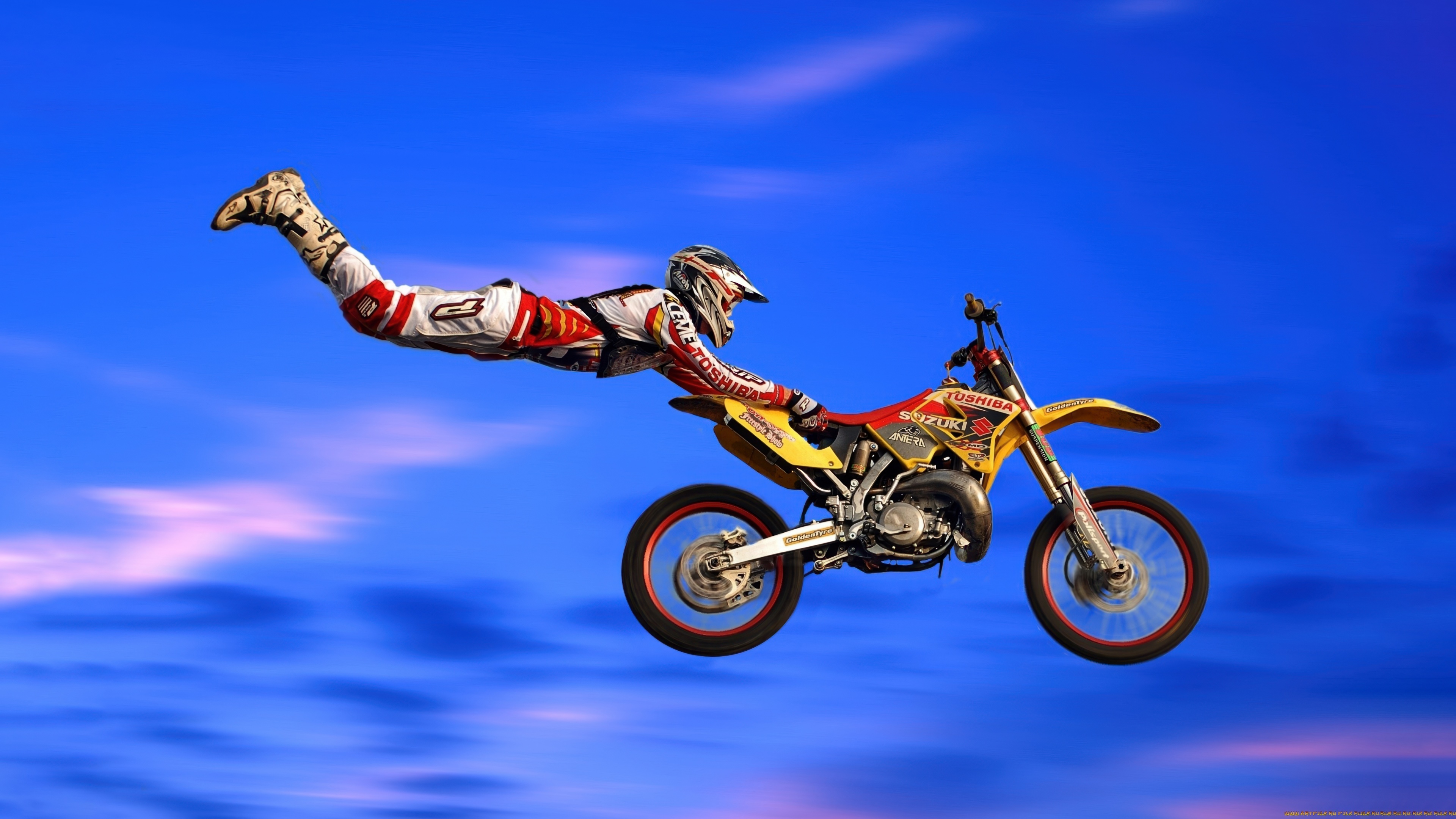 Motocross rider performing a mid-air stunt on a blue Suzuki RM motorcycle against a bright blue sky.