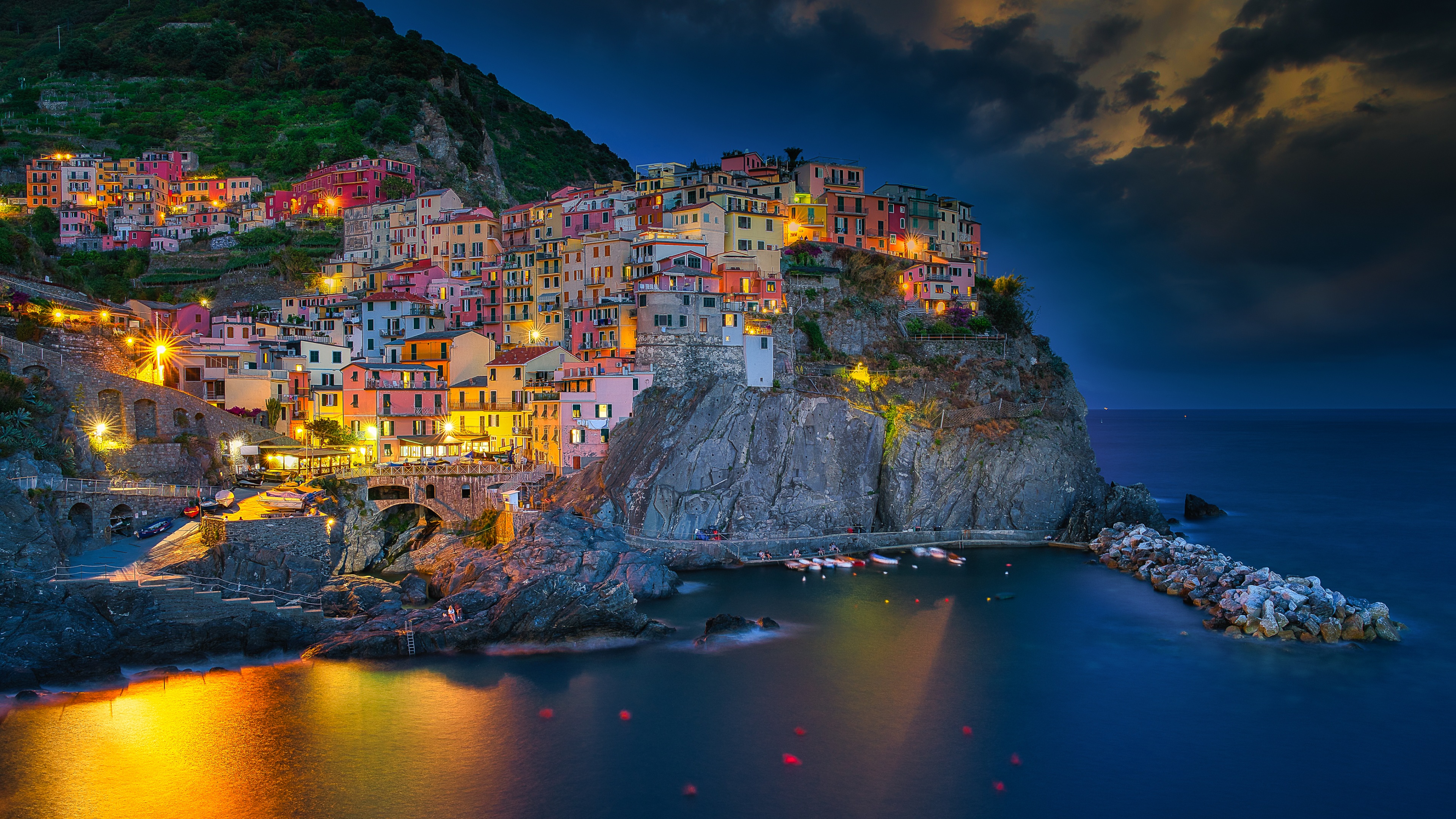 A long exposure view of Manarola village in Cinque Terre, Italy, during blue hour with glowing night lights reflecting on the ocean.