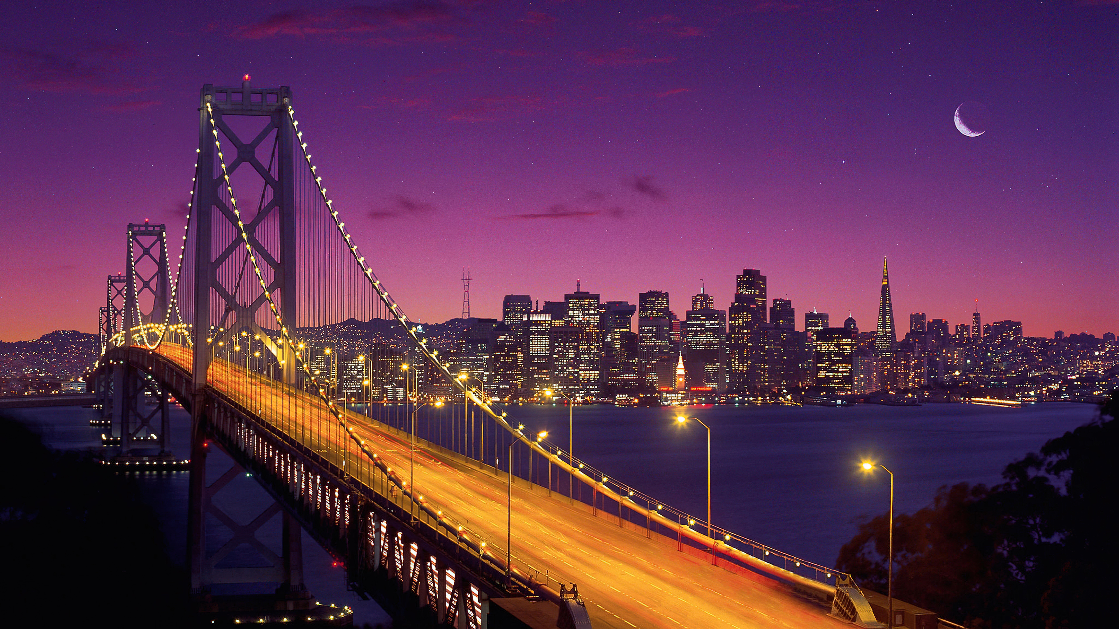 A long exposure photograph of the San Francisco-Oakland Bay Bridge under a purple twilight sky with a crescent moon.