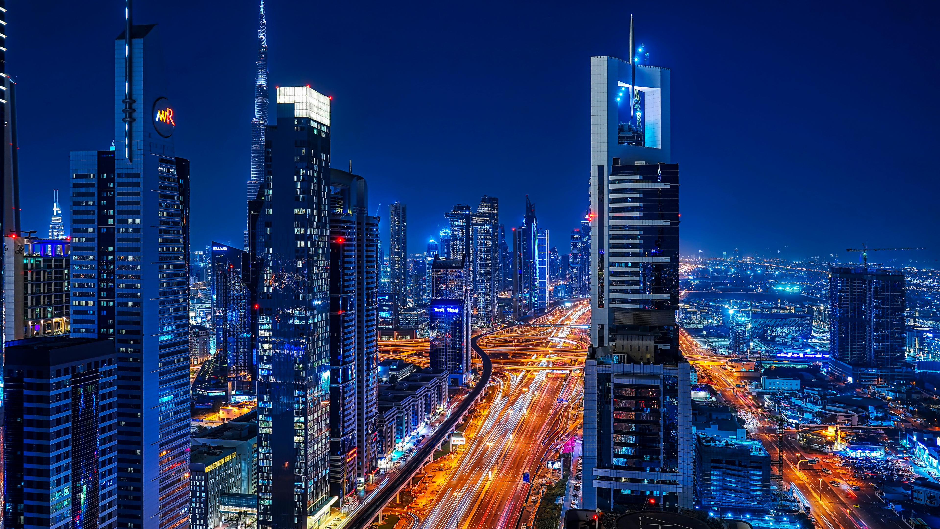 A long exposure night view of the Dubai skyline featuring illuminated skyscrapers and city lights along Sheikh Zayed Road.