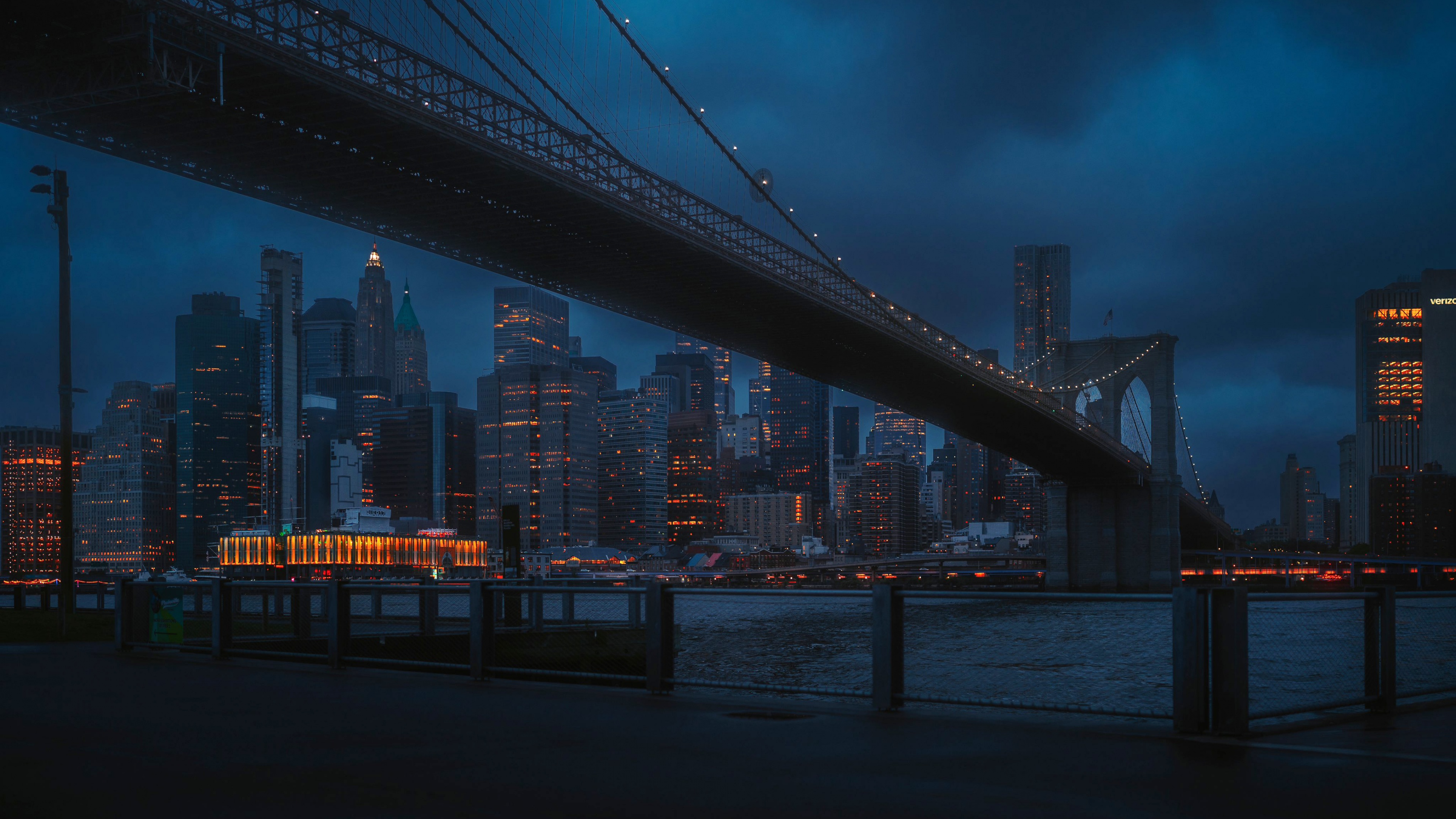 A dark and atmospheric night view of the Brooklyn Bridge in New York City, showcasing the illuminated bridge structure and distant city lights.