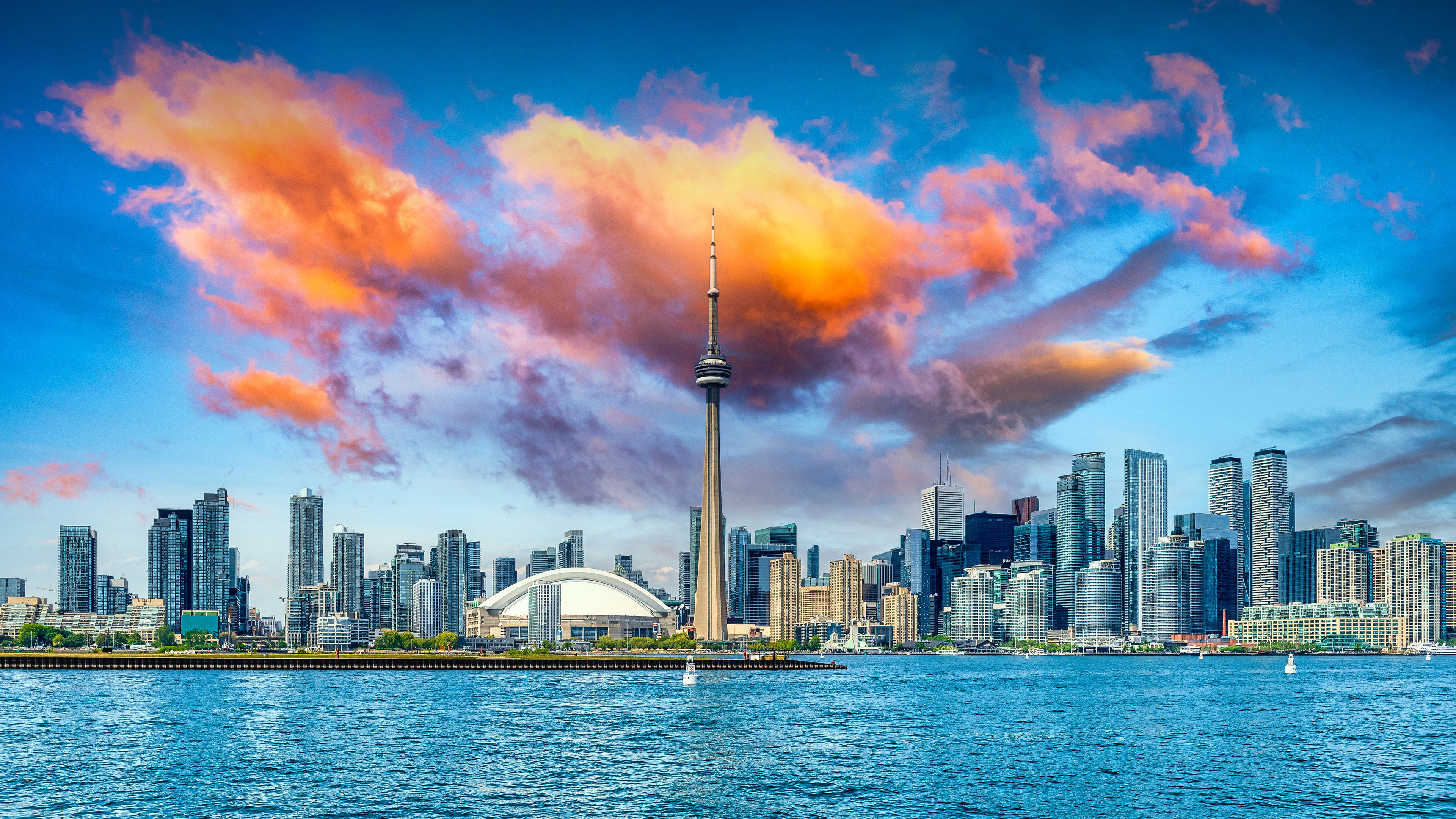 A panoramic view of the Toronto skyline, featuring the CN Tower and Lake Ontario under a cloudy sky.