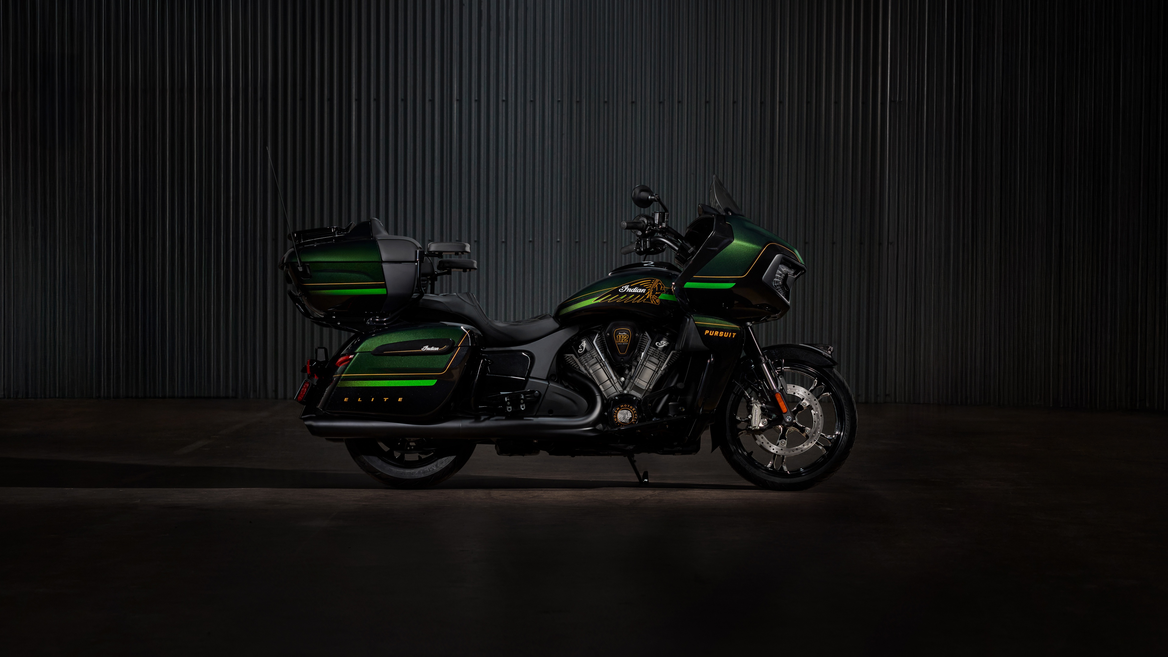 A cinematic close-up of a 2025 Indian Pursuit Elite motorcycle with a dark green finish against a dark, atmospheric background.