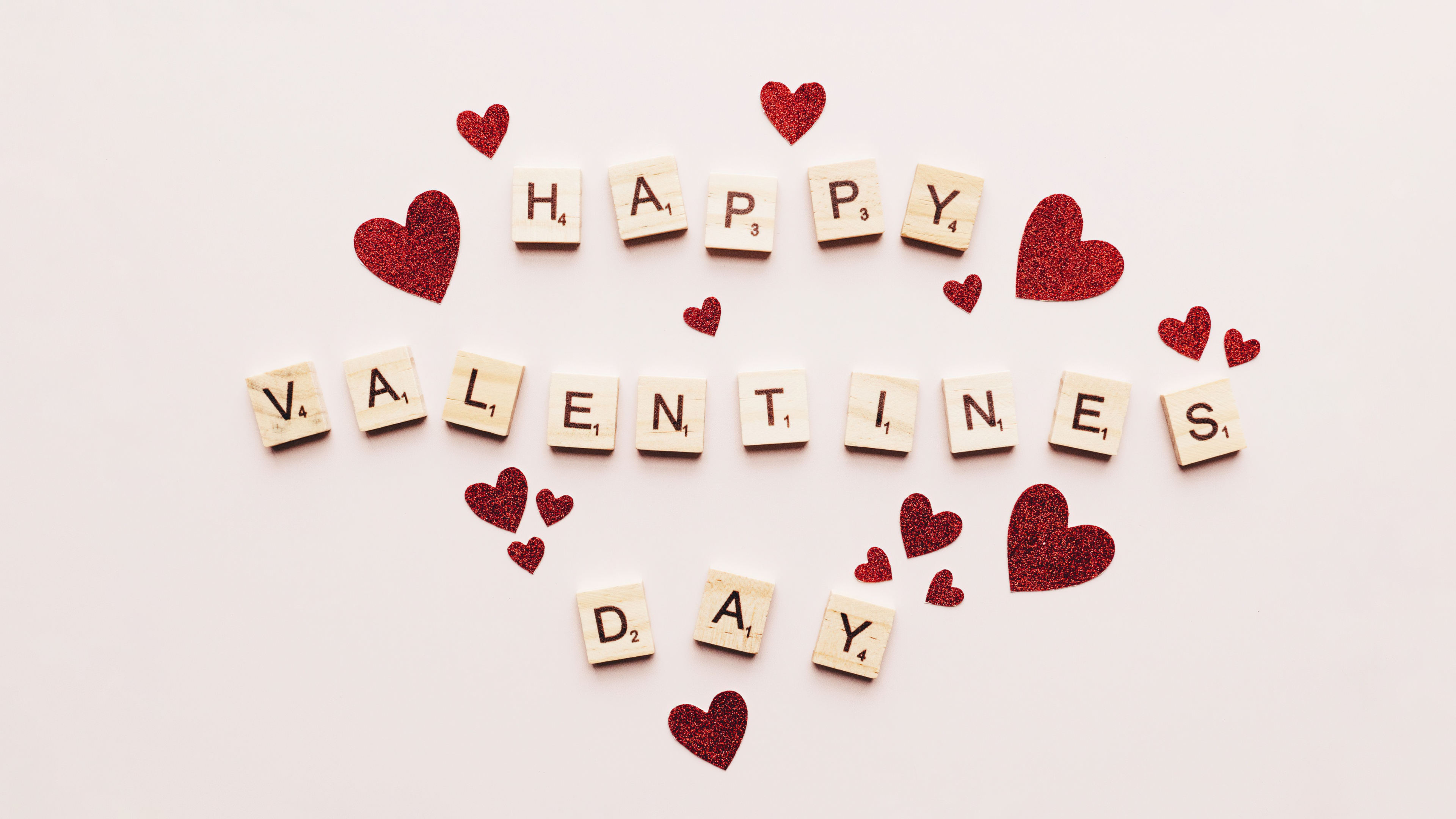 Wooden Scrabble tiles spelling 'Happy Valentine's Day' decorated with small red hearts on a plain white background.
