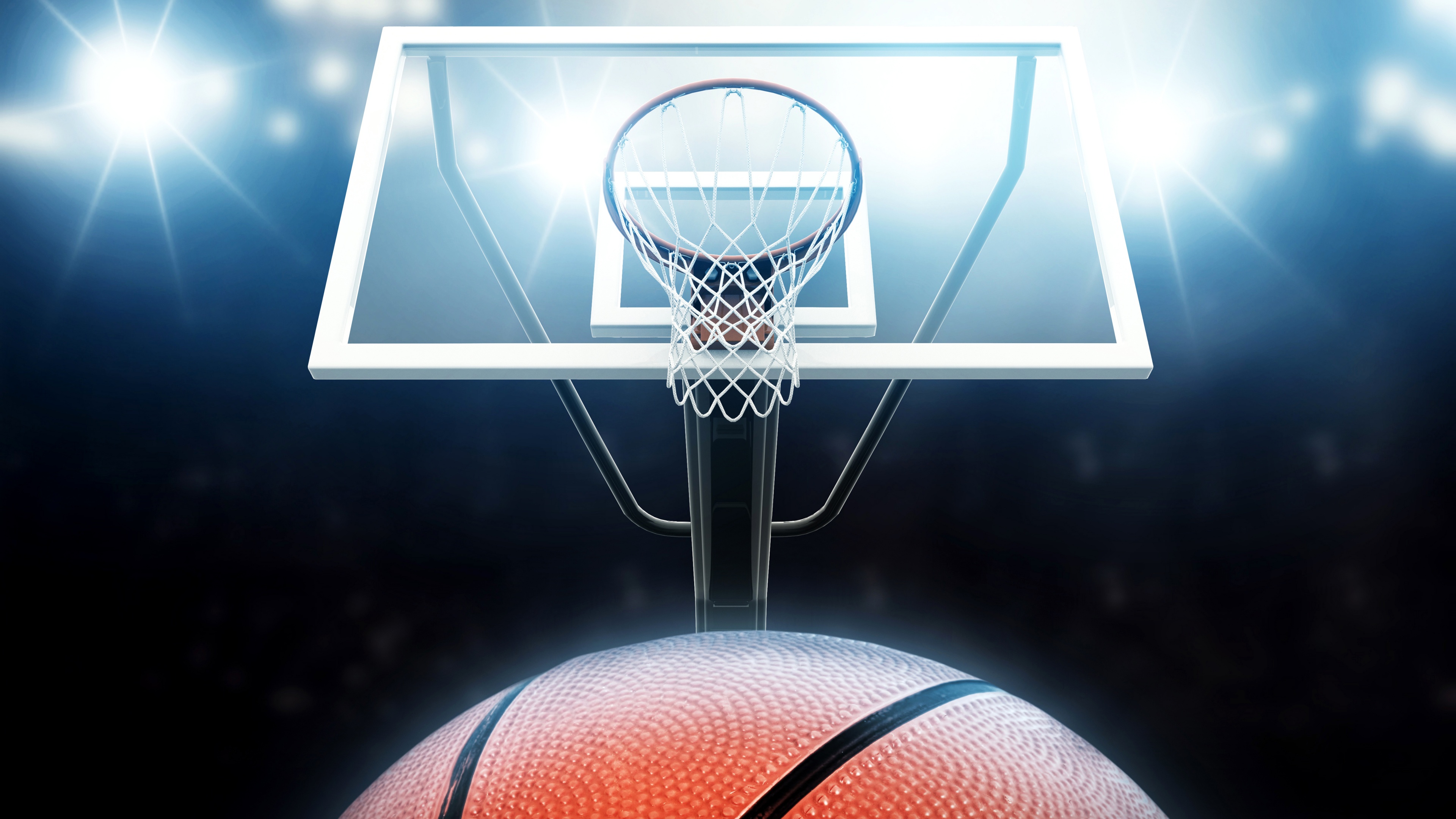 A close-up shot of a basketball hoop and backboard illuminated by dramatic lighting on a basketball court.