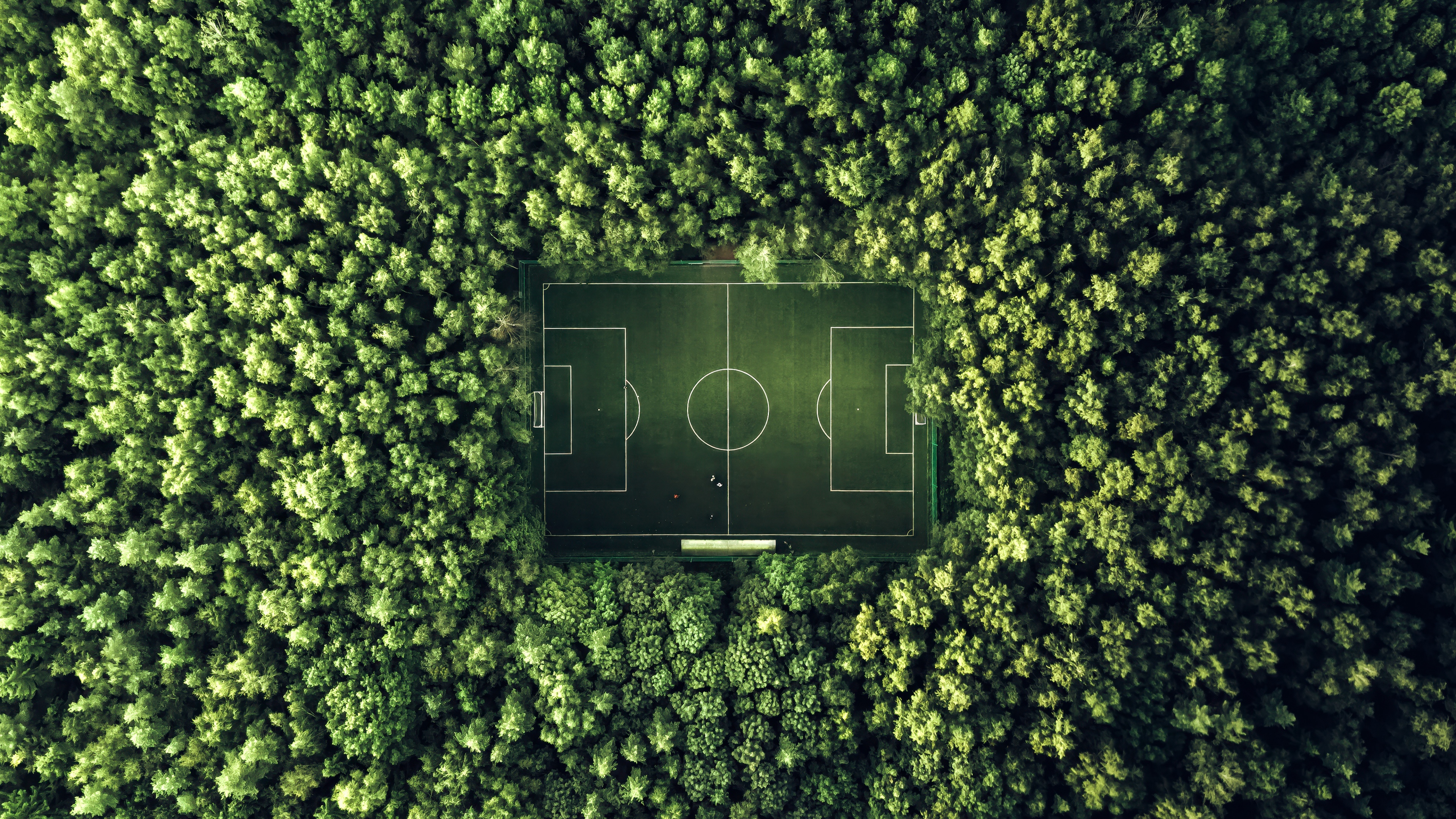 An aerial drone view of a symmetrical soccer field surrounded by a dense, lush green forest and wilderness.