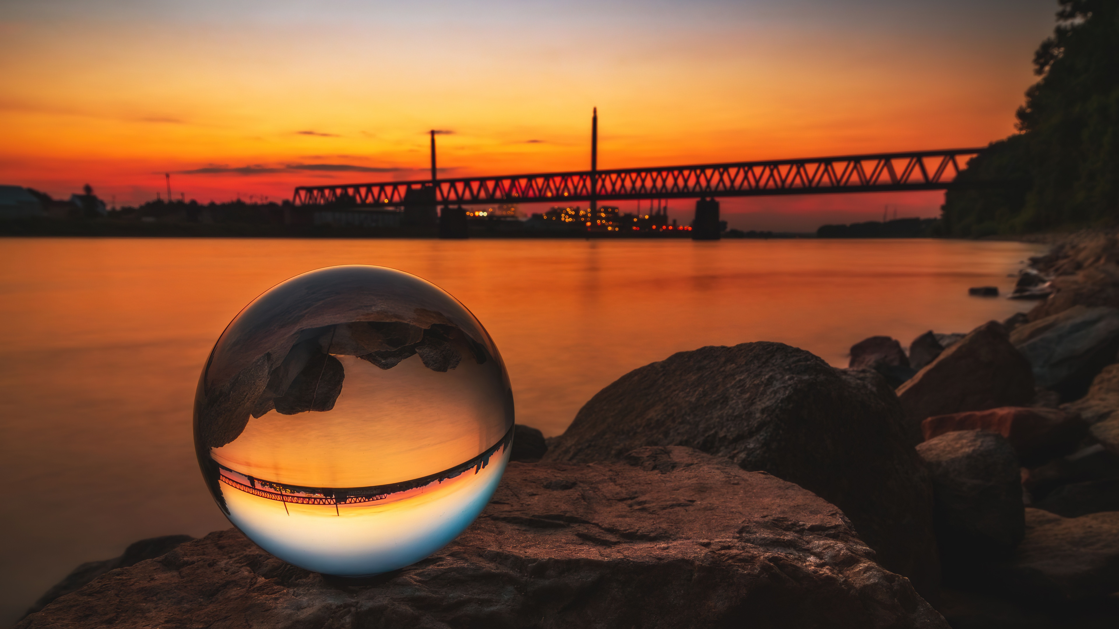 A close-up macro shot of a glowing crystal ball resting on textured stones near a bridge during a vibrant sunset.