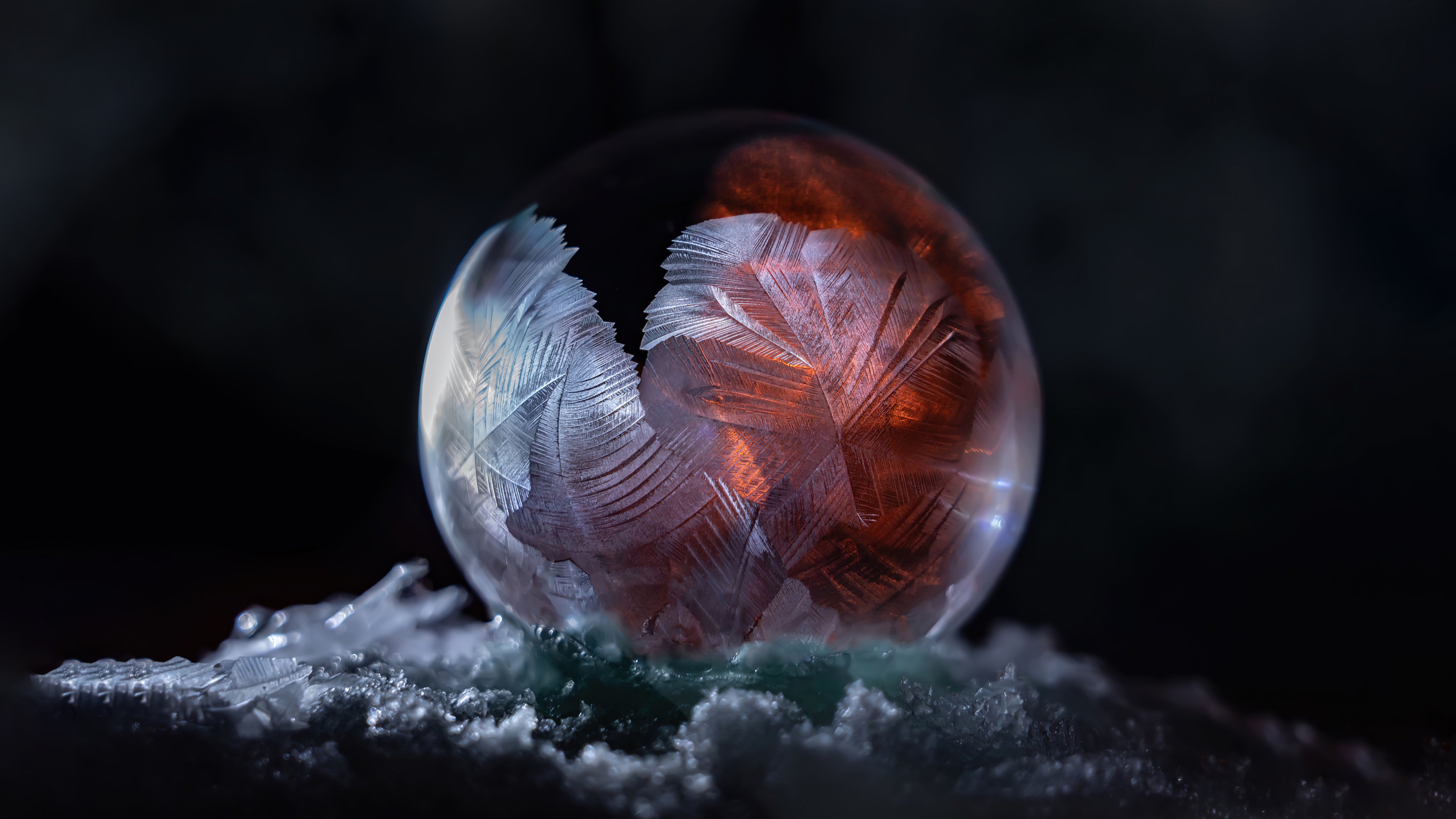 A close-up macro photograph of an iridescent soap bubble resting among white snowflakes in a dark, wintry setting.