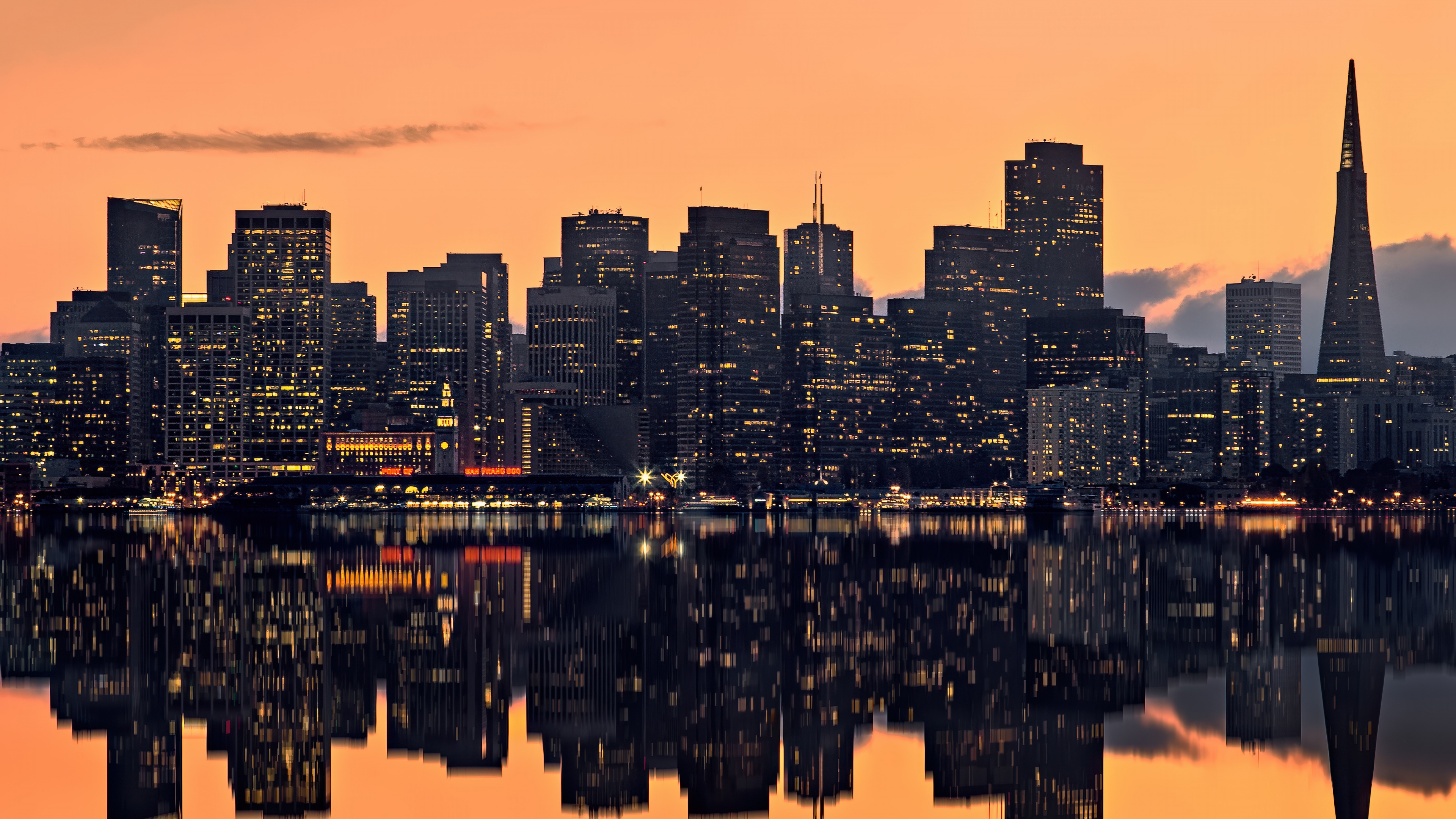 A panoramic view of the San Francisco skyline at sunset with a vibrant orange sky and reflections on high-rise buildings.