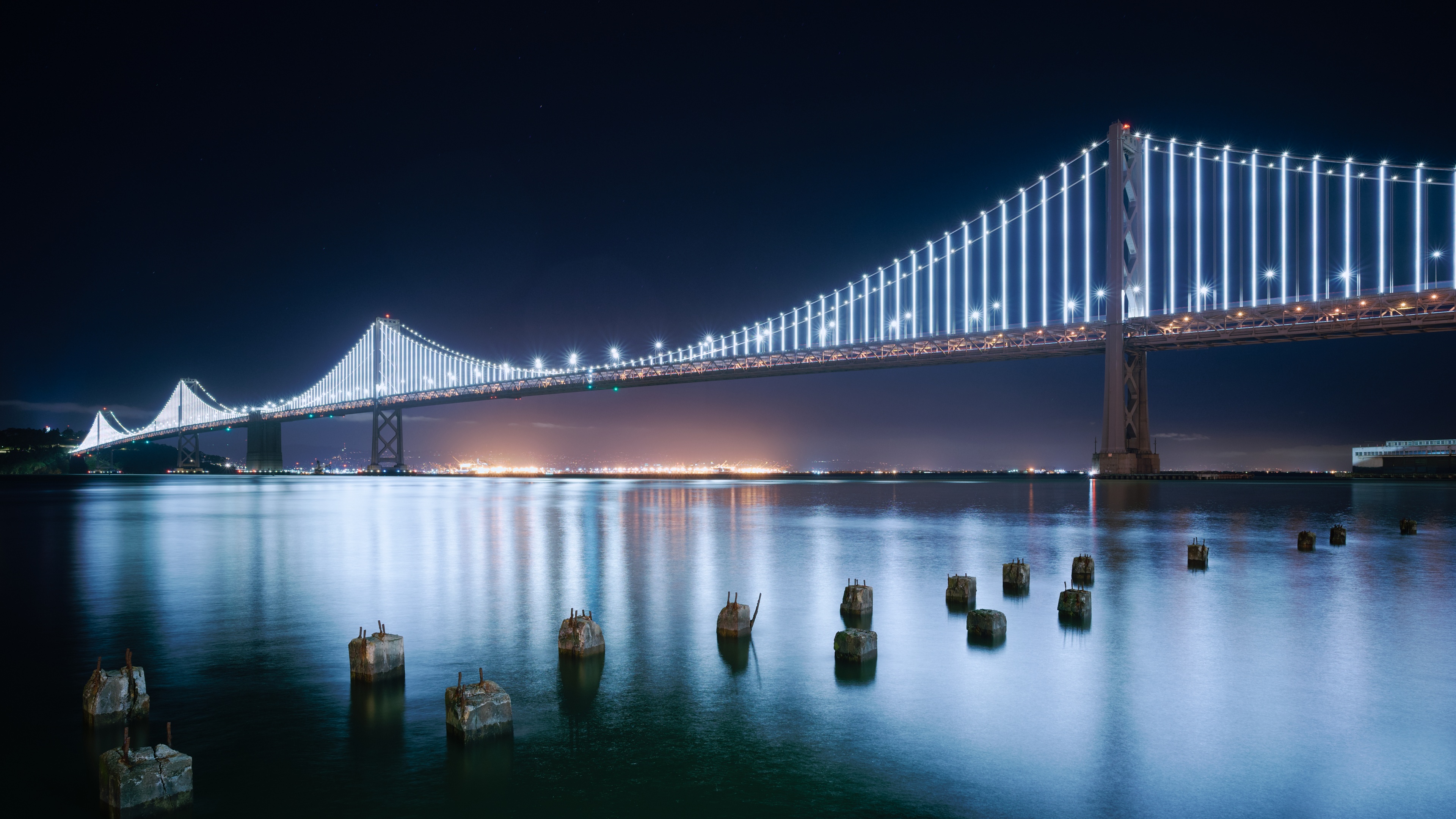 San Francisco-Oakland Bay Bridge at night with bright lights reflecting on the water surface.