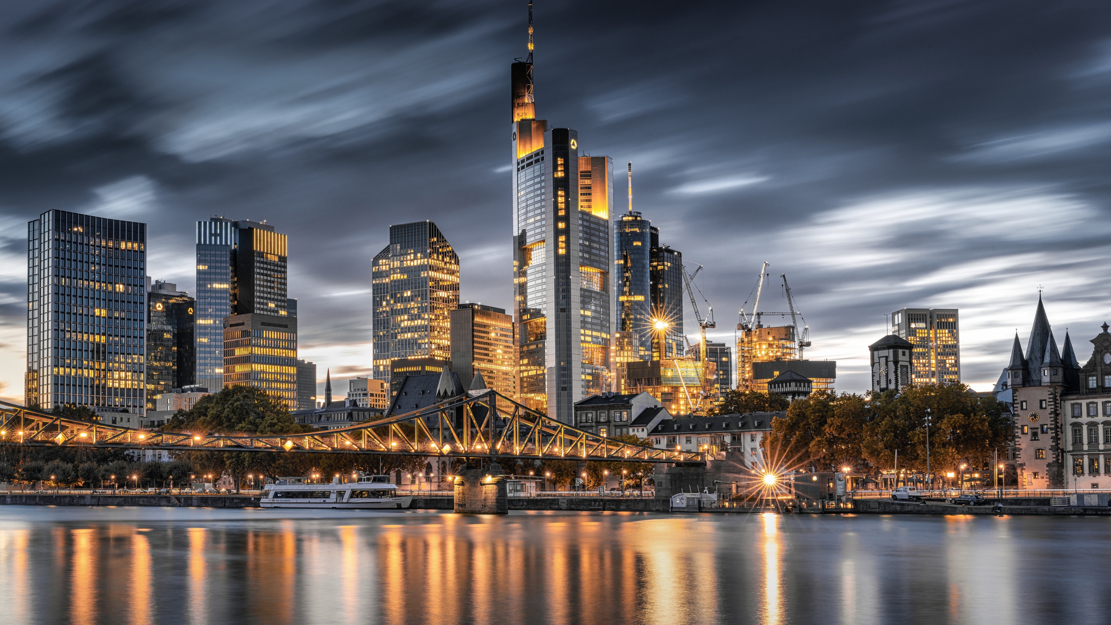 A high-resolution night view of the Frankfurt, Germany city skyline with bright skyscraper lights reflecting on the river.