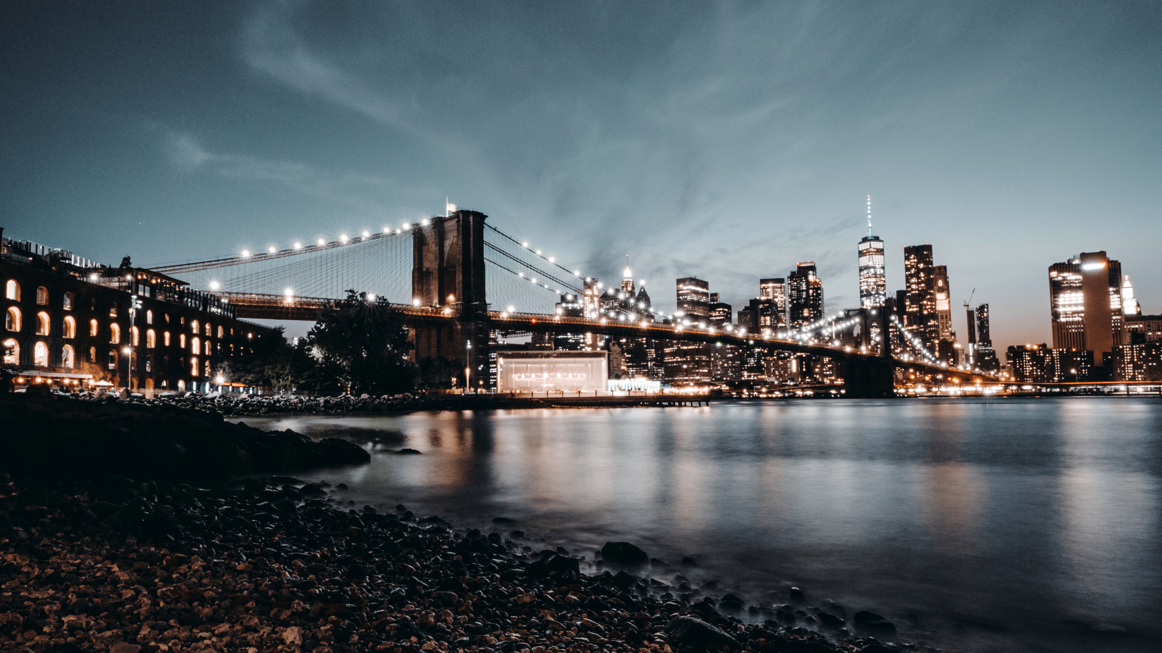 A nighttime view of the Brooklyn Bridge in New York City featuring a crescent moon and city lights reflecting on the water.