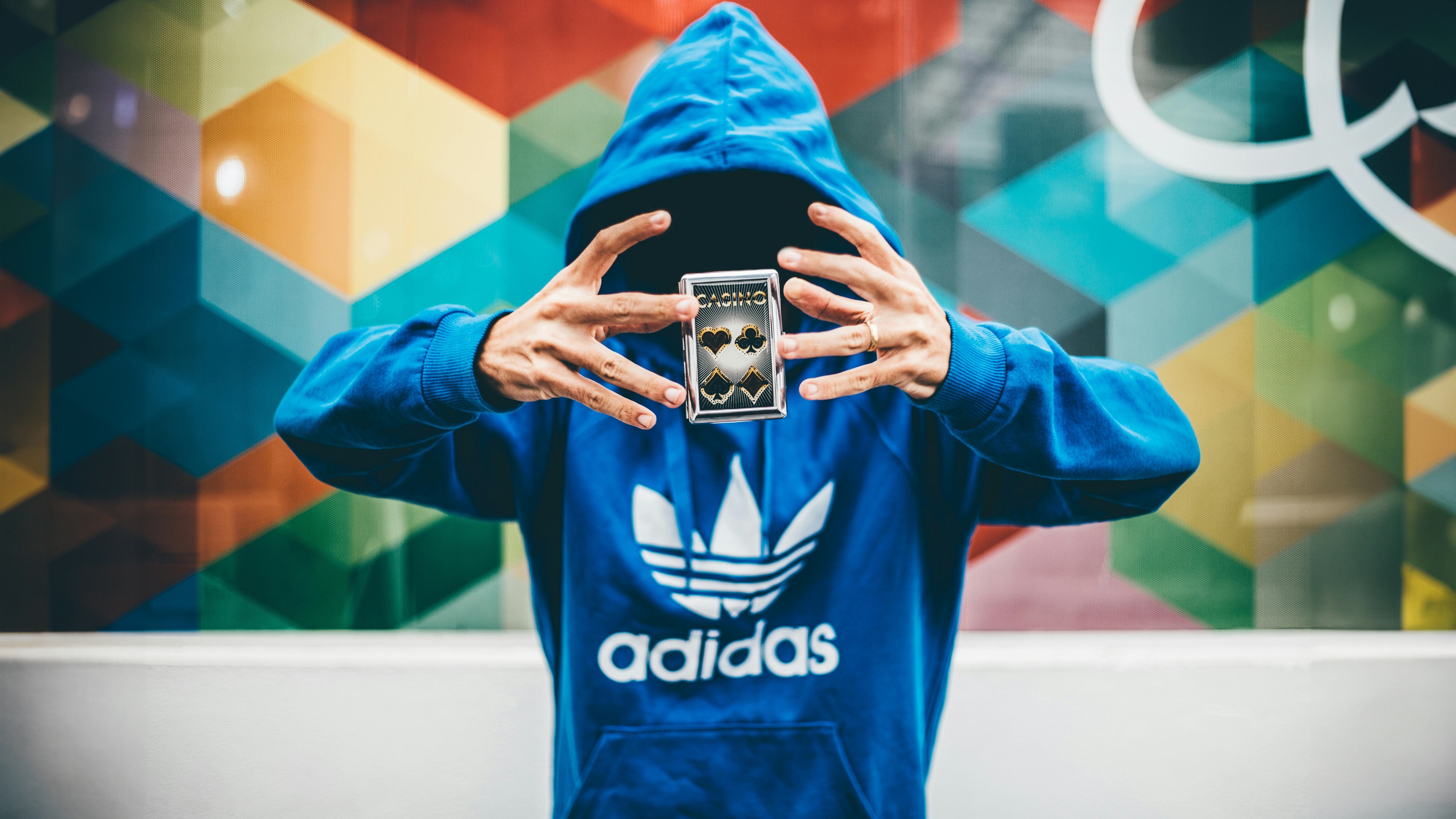 A man wearing an Adidas hoodie surrounded by playing cards in a dark casino-themed setting.