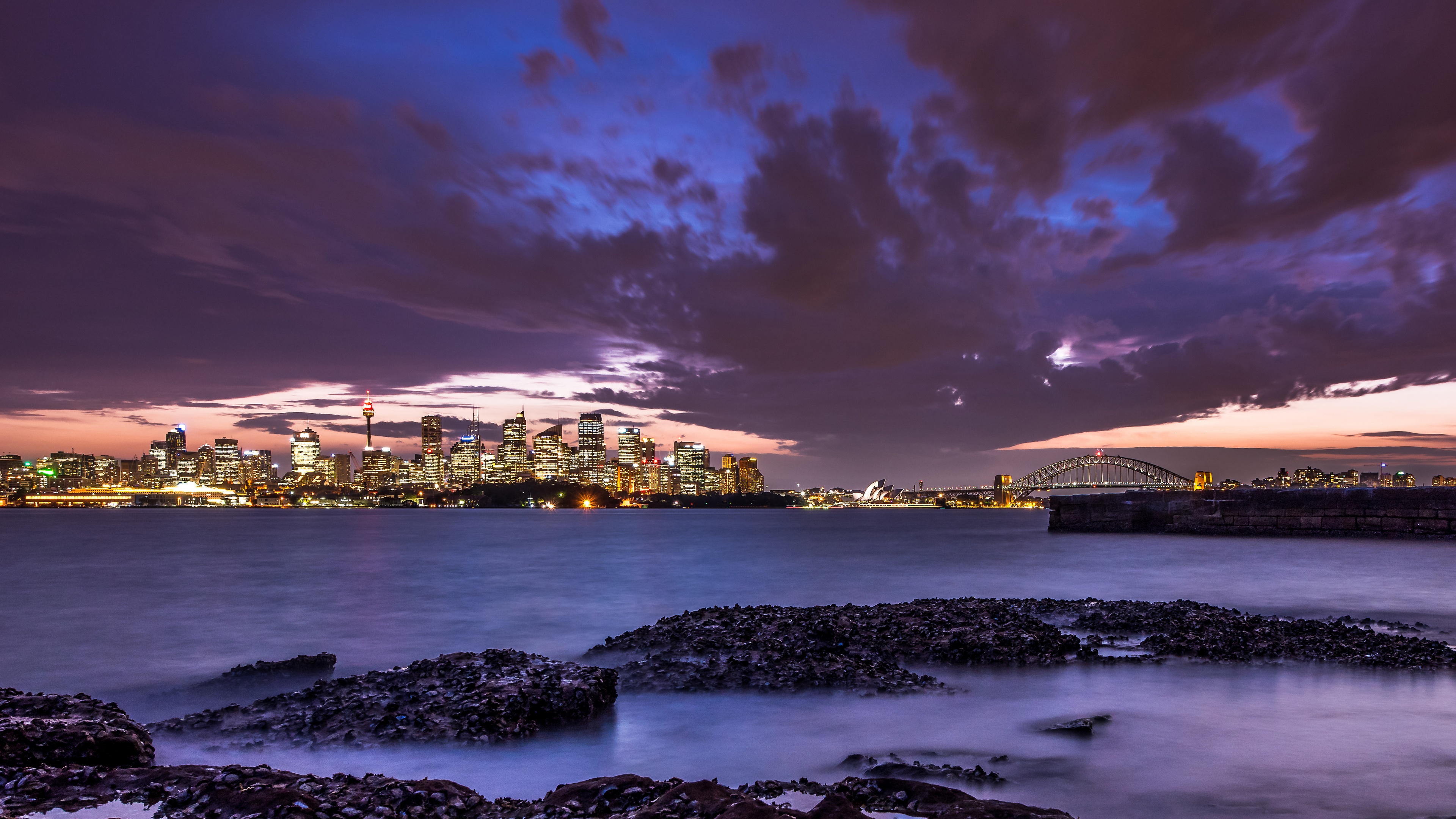 A scenic dusk view of the Sydney, Australia skyline and harbour with glowing city lights.