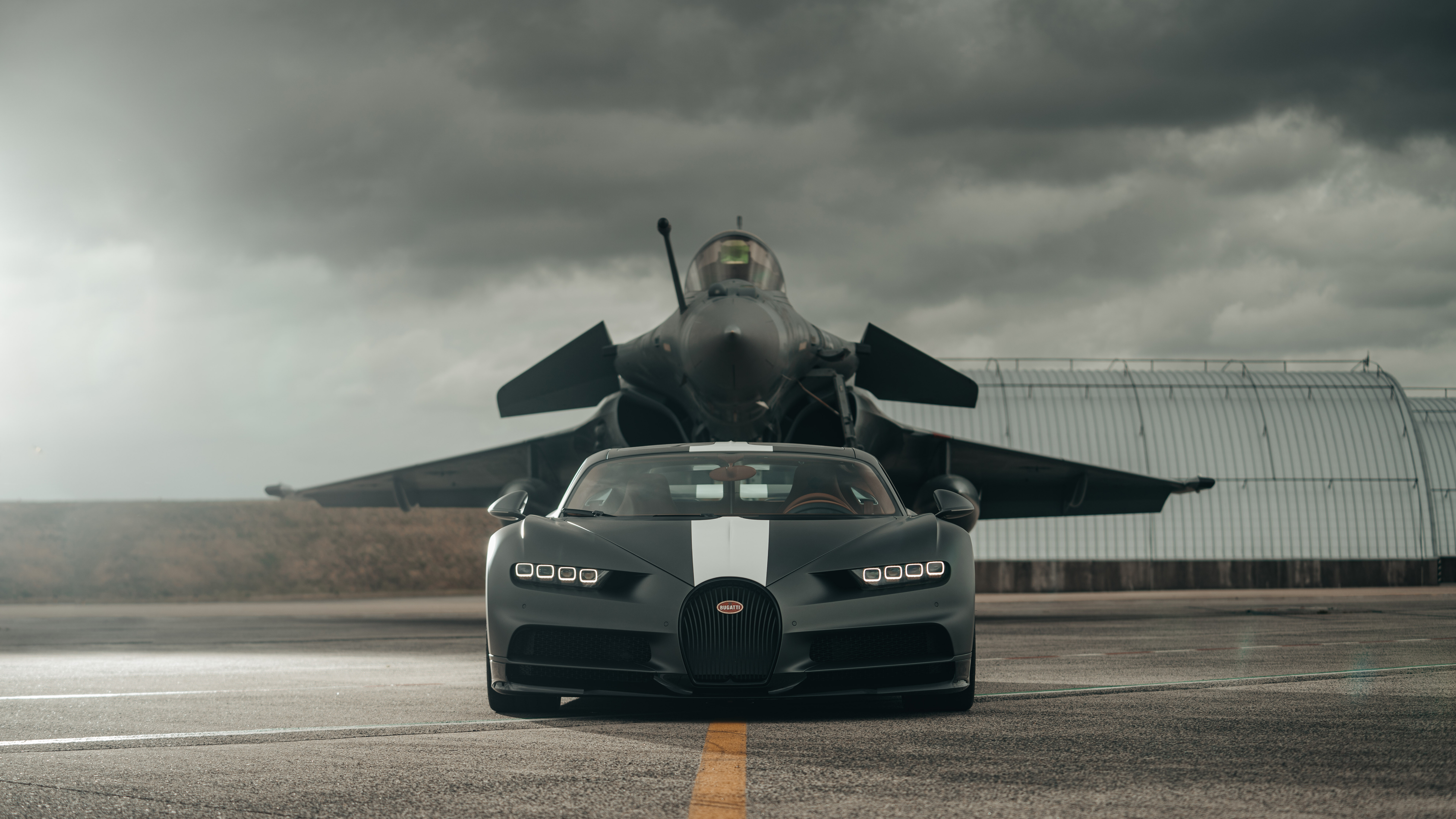 A black Bugatti Chiron supercar parked next to a stealth jet fighter in a dark, atmospheric setting.