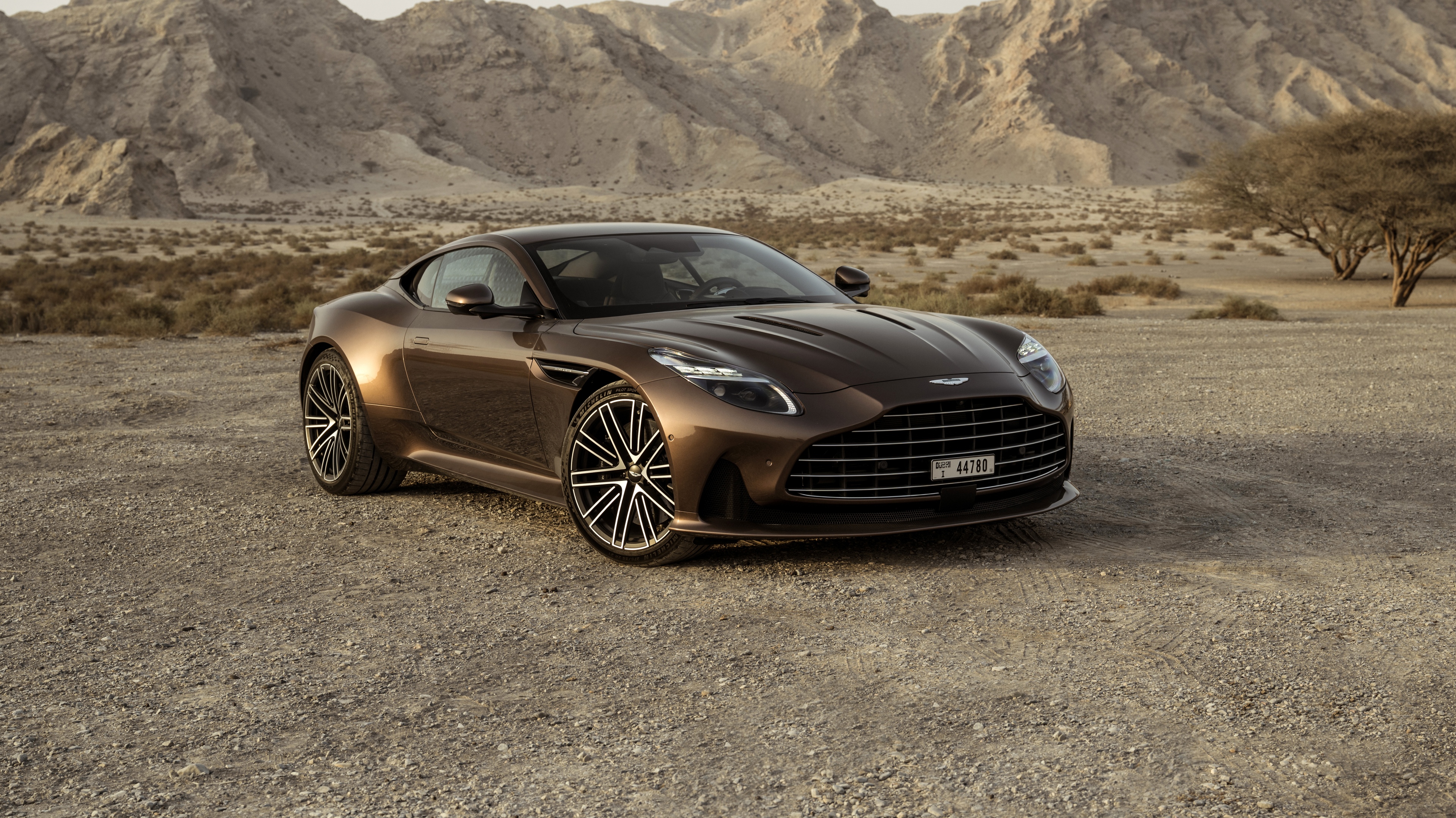 A high-resolution shot of an Aston Martin DB12 driving through golden desert sand dunes.