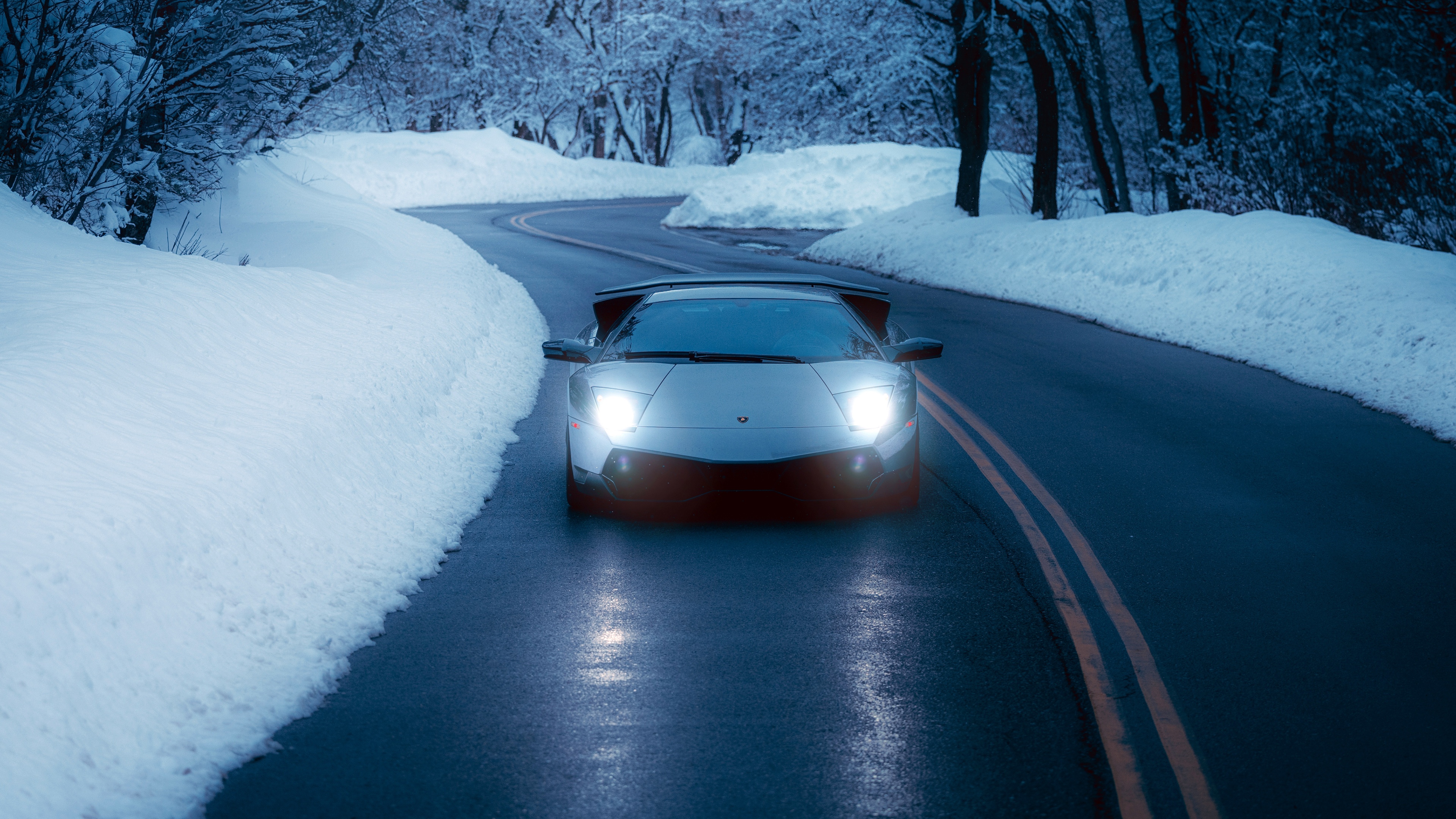 A Lamborghini Murcielago driving on a snow-covered road at night with bright headlights and a cinematic blue aesthetic.