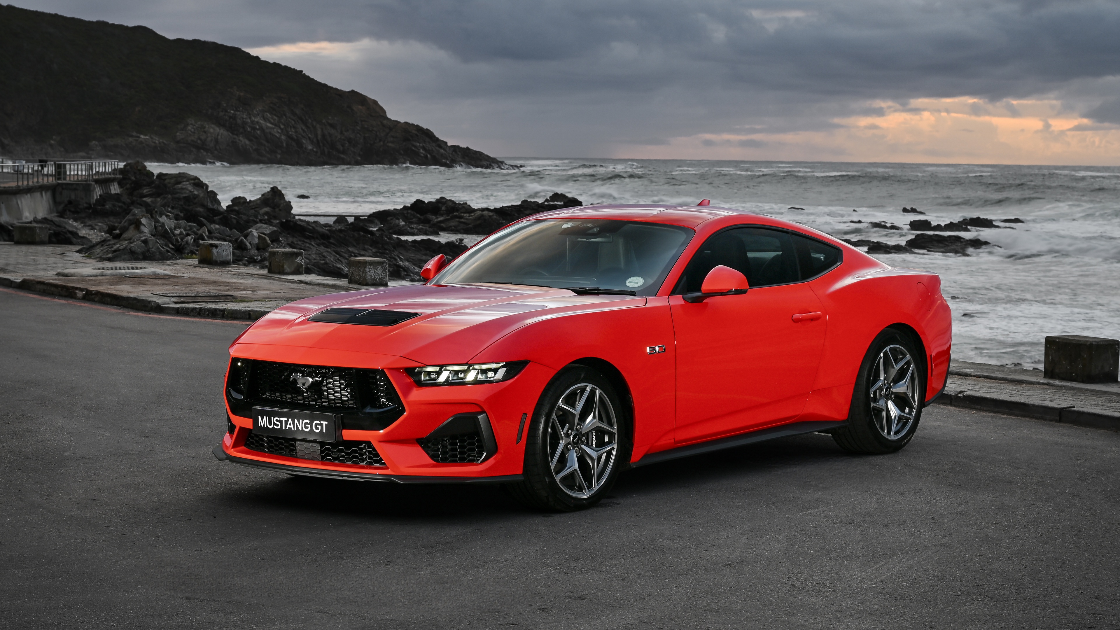 A vibrant red 2024 Ford Mustang GT parked on the sand with ocean waves in the background.