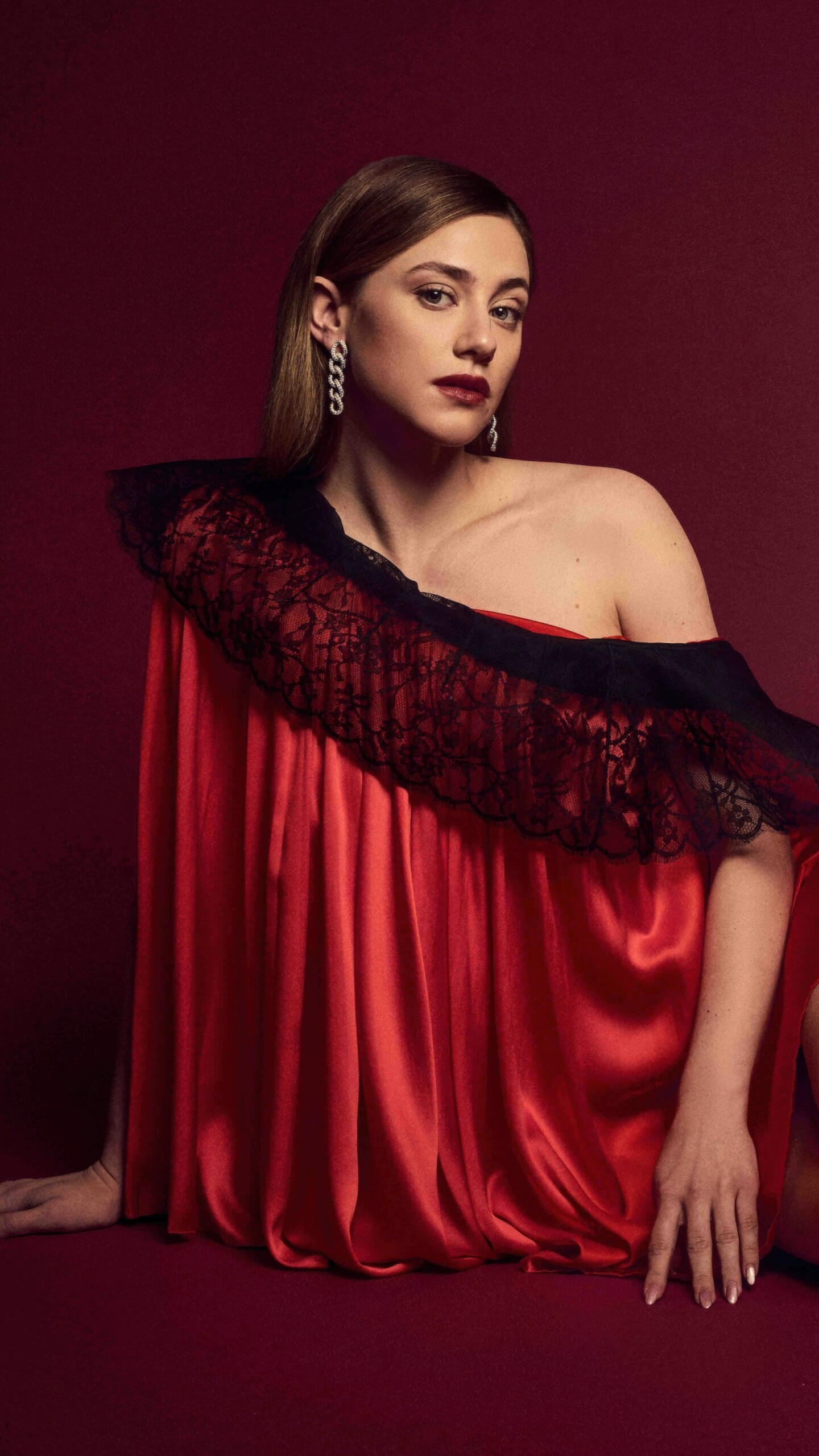 A high-fashion portrait of Lili Reinhart wearing an elegant red dress against a vibrant red background.