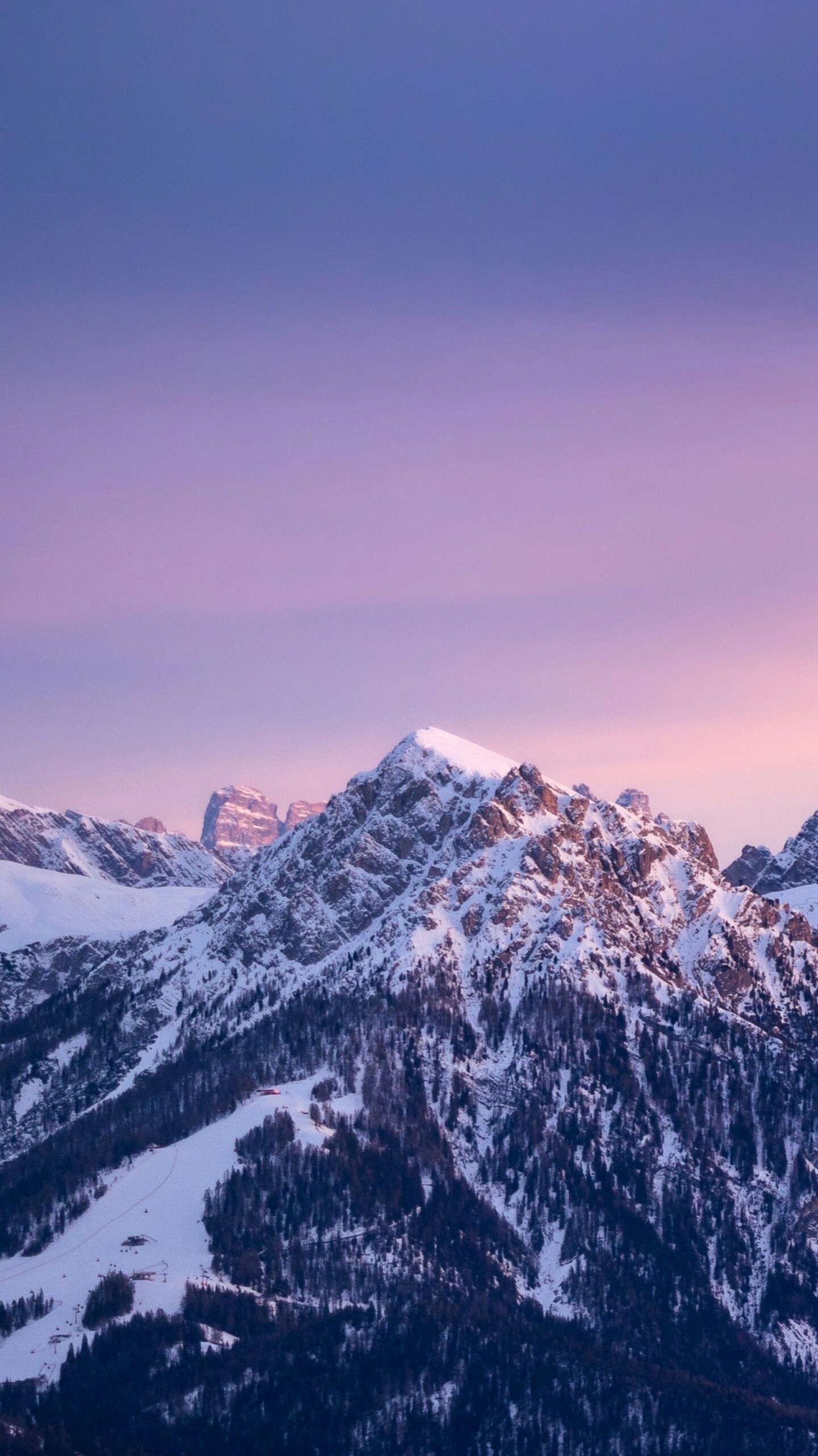 A wide panoramic view of snow-covered mountain peaks under a colorful gradient sky during a golden hour sunset.