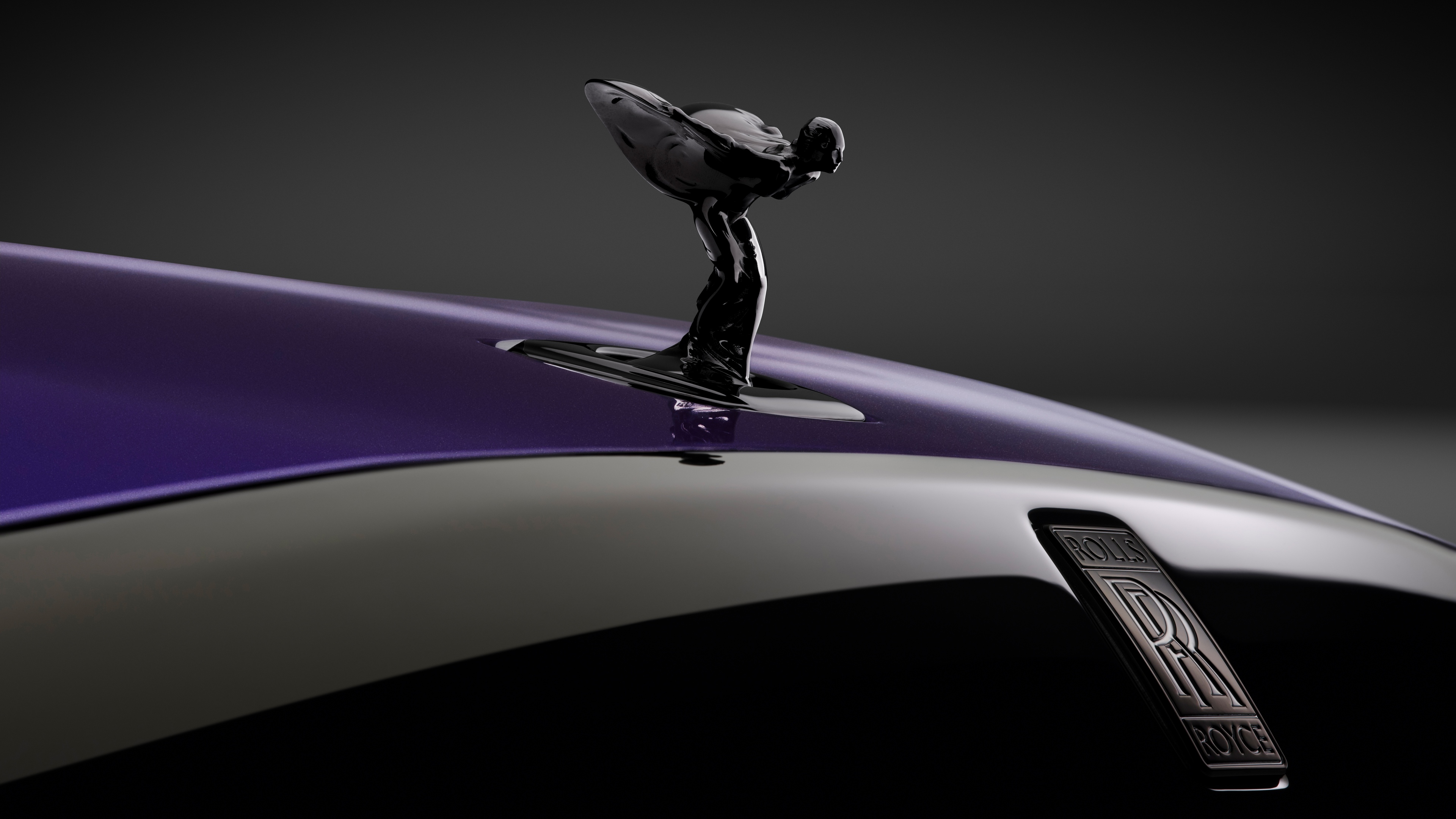 A detailed close-up of the Spirit of Ecstasy ornament on the hood of a 2025 Rolls-Royce Black Badge Spectre against a dark, cinematic background.
