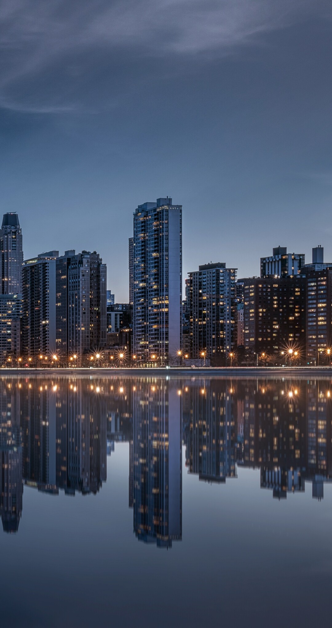 The illuminated Chicago skyline and skyscrapers reflecting on the water under a dramatic evening sky at night.
