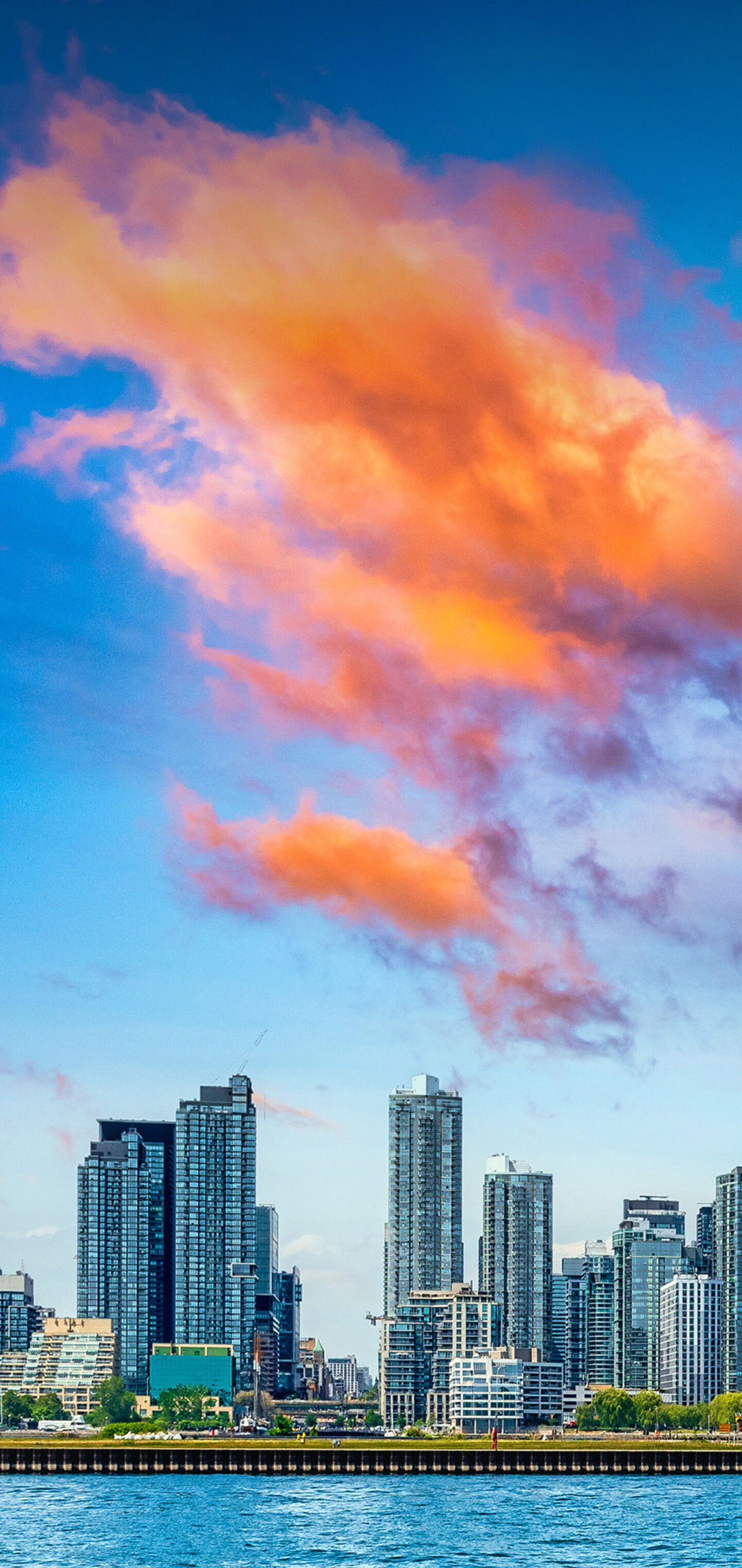 A panoramic cityscape of Toronto, Canada, featuring the CN Tower and the urban skyline under a moody, cloudy sky overlooking Lake Ontario.
