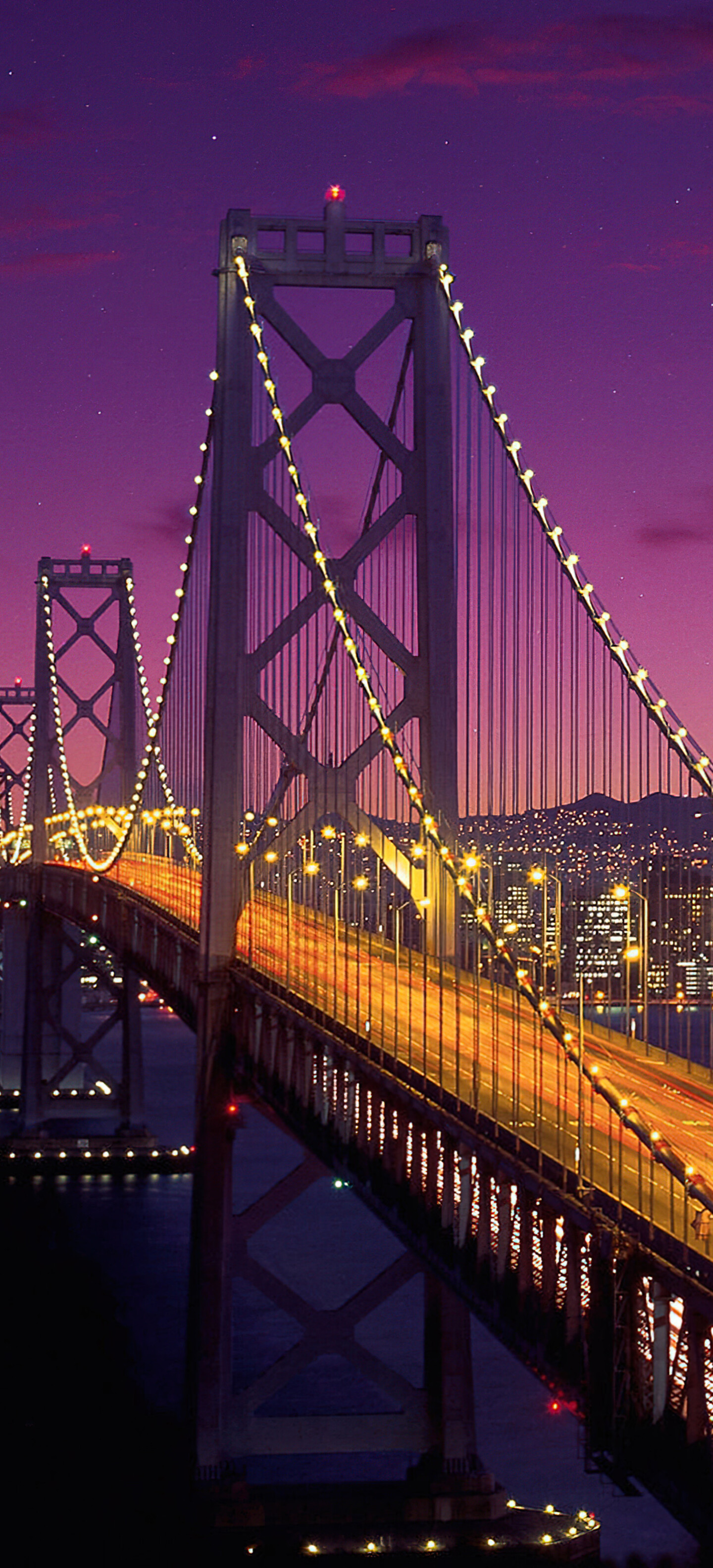 A long exposure photograph of the San Francisco-Oakland Bay Bridge under a deep purple twilight sky with a crescent moon.