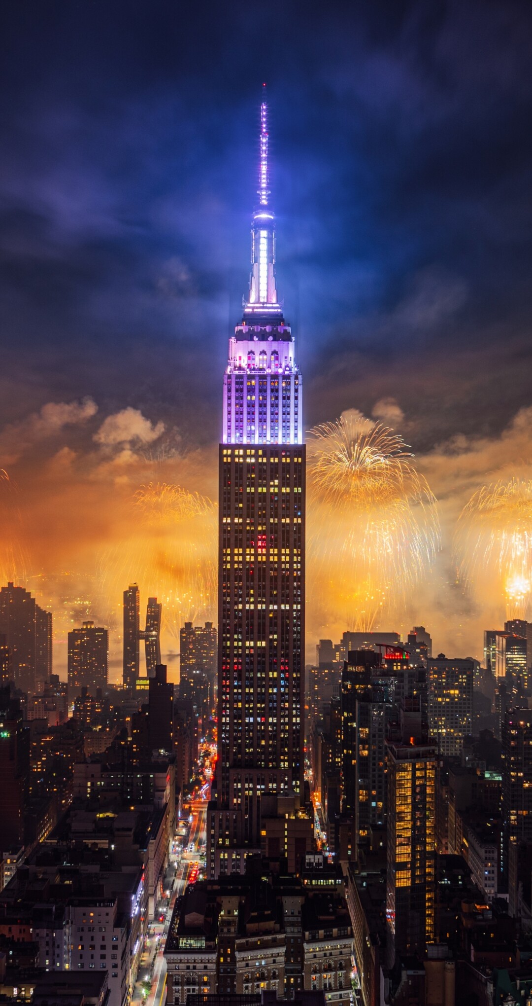 A night view of the Empire State Building in New York City with colorful fireworks exploding in the sky over skyscrapers.