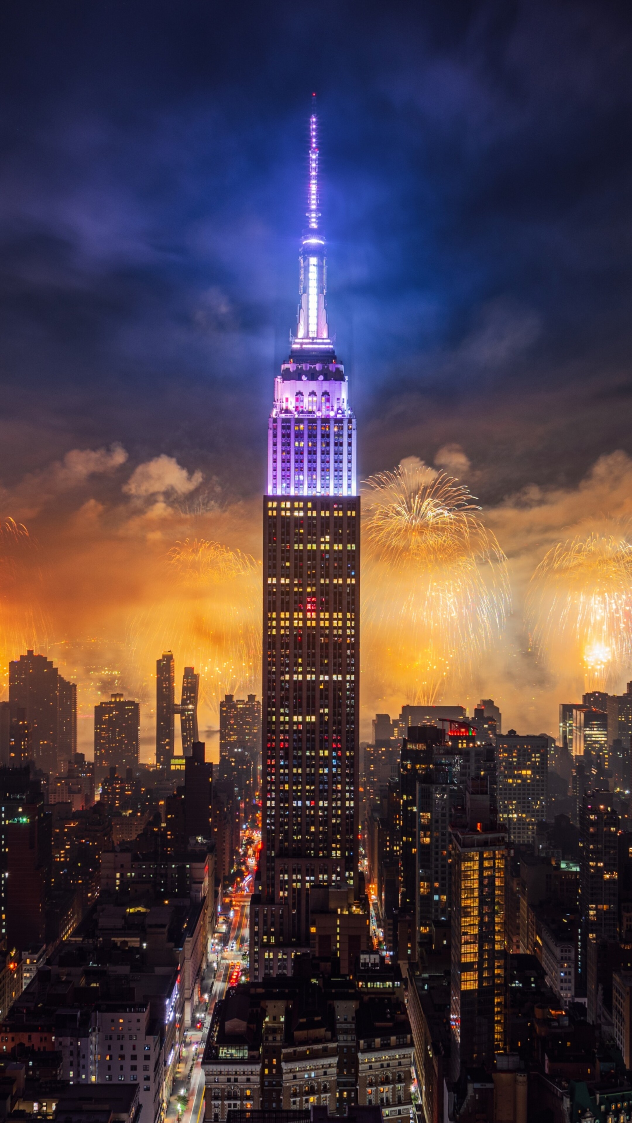 A night view of the Empire State Building in New York City with colorful fireworks exploding in the sky over skyscrapers.