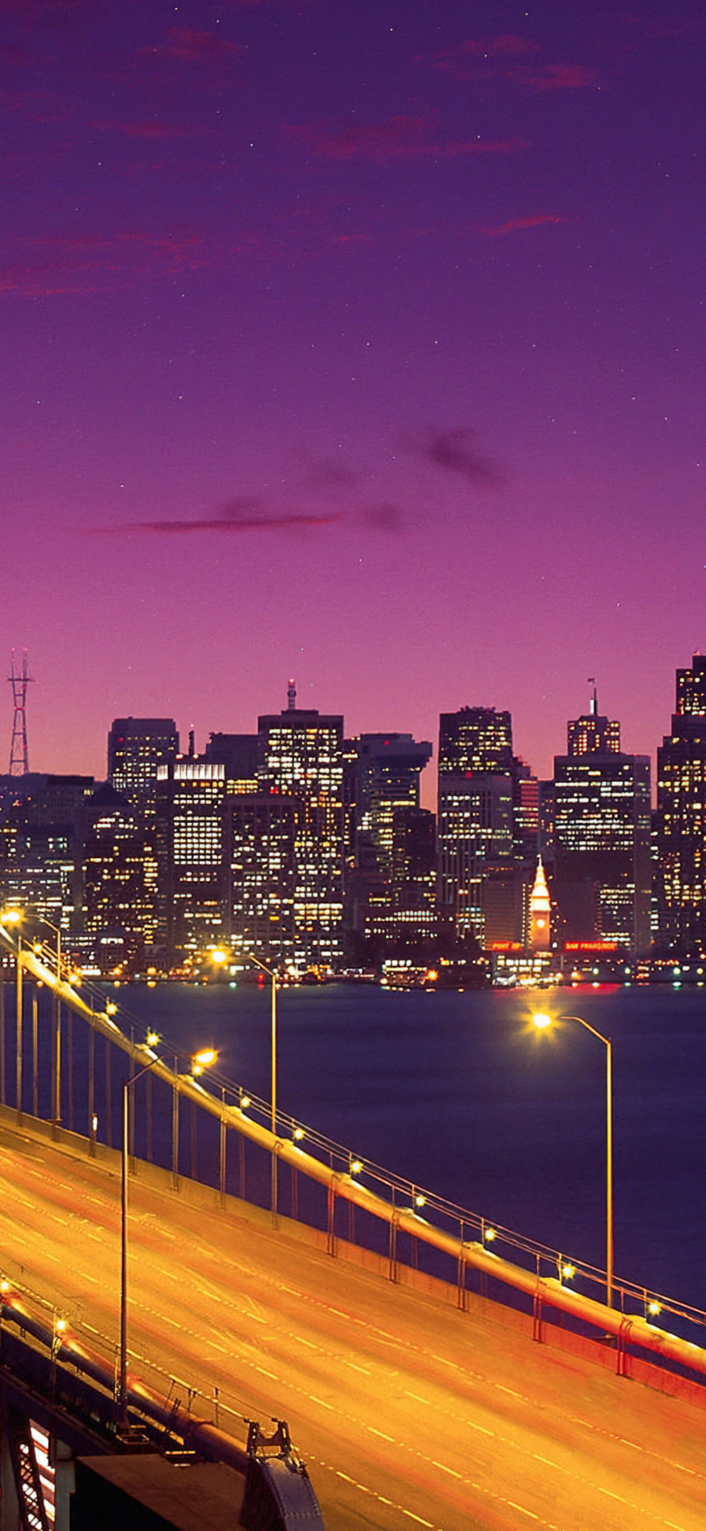 A long exposure shot of the San Francisco-Oakland Bay Bridge under a purple twilight sky with a crescent moon.