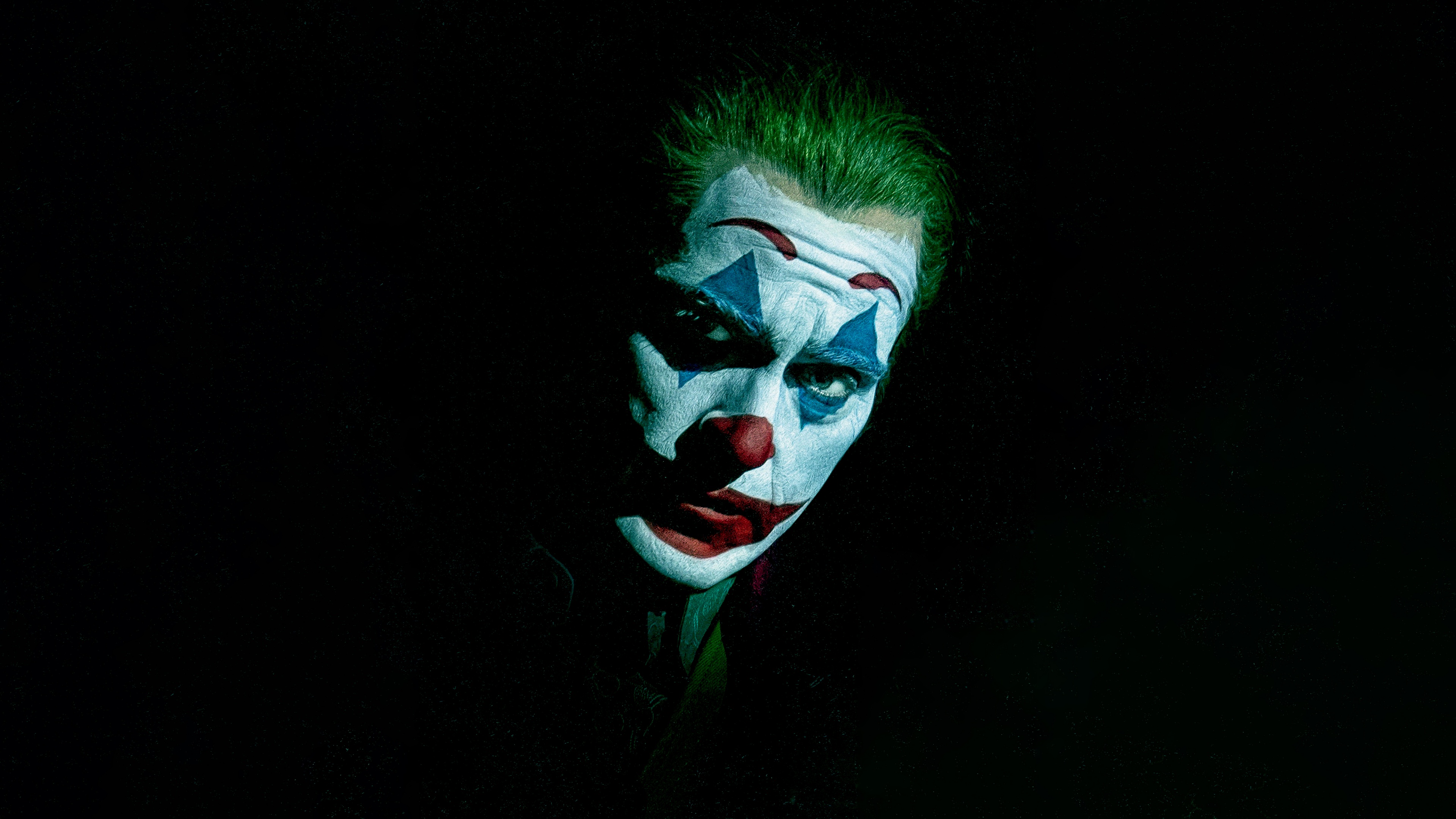 A dramatic close-up portrait of Joaquin Phoenix as the Joker in Joker: Folie à Deux against a dark background.