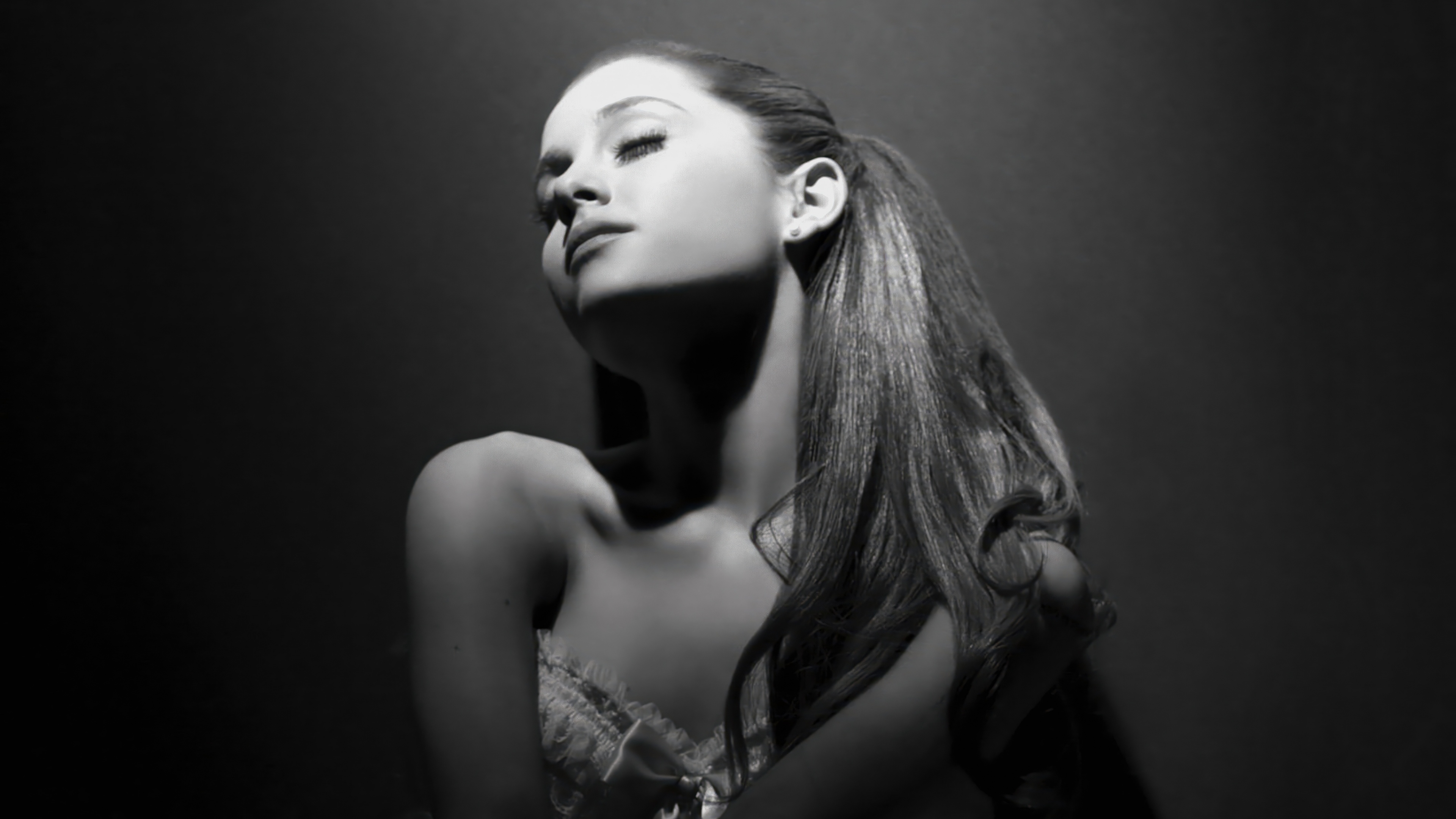 A high-contrast, black and white close-up portrait of Ariana Grande with a dark, moody background.