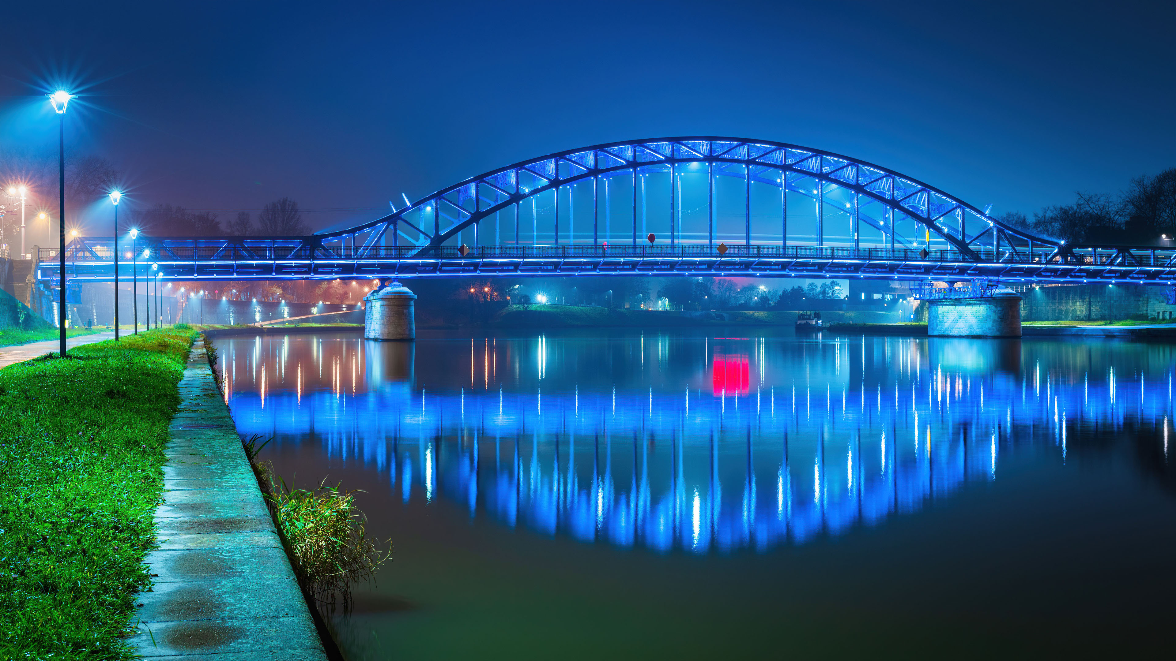 A scenic night view of the illuminated Józef Piłsudski Bridge in Kraków, Poland, with city lights reflecting on the river.