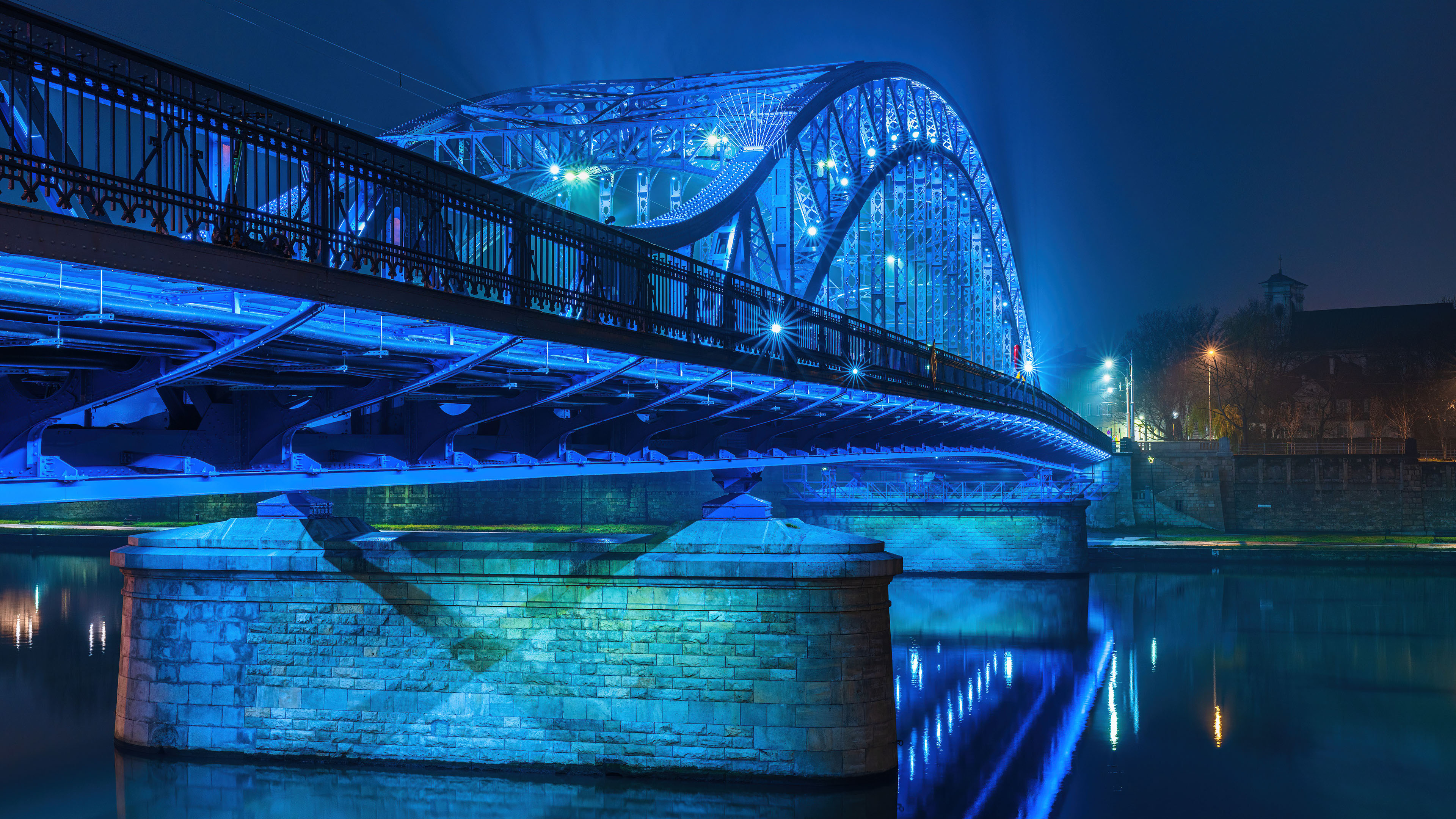 Night view of the illuminated Józef Piłsudski Bridge in Kraków, Poland, with city lights reflecting on the water.