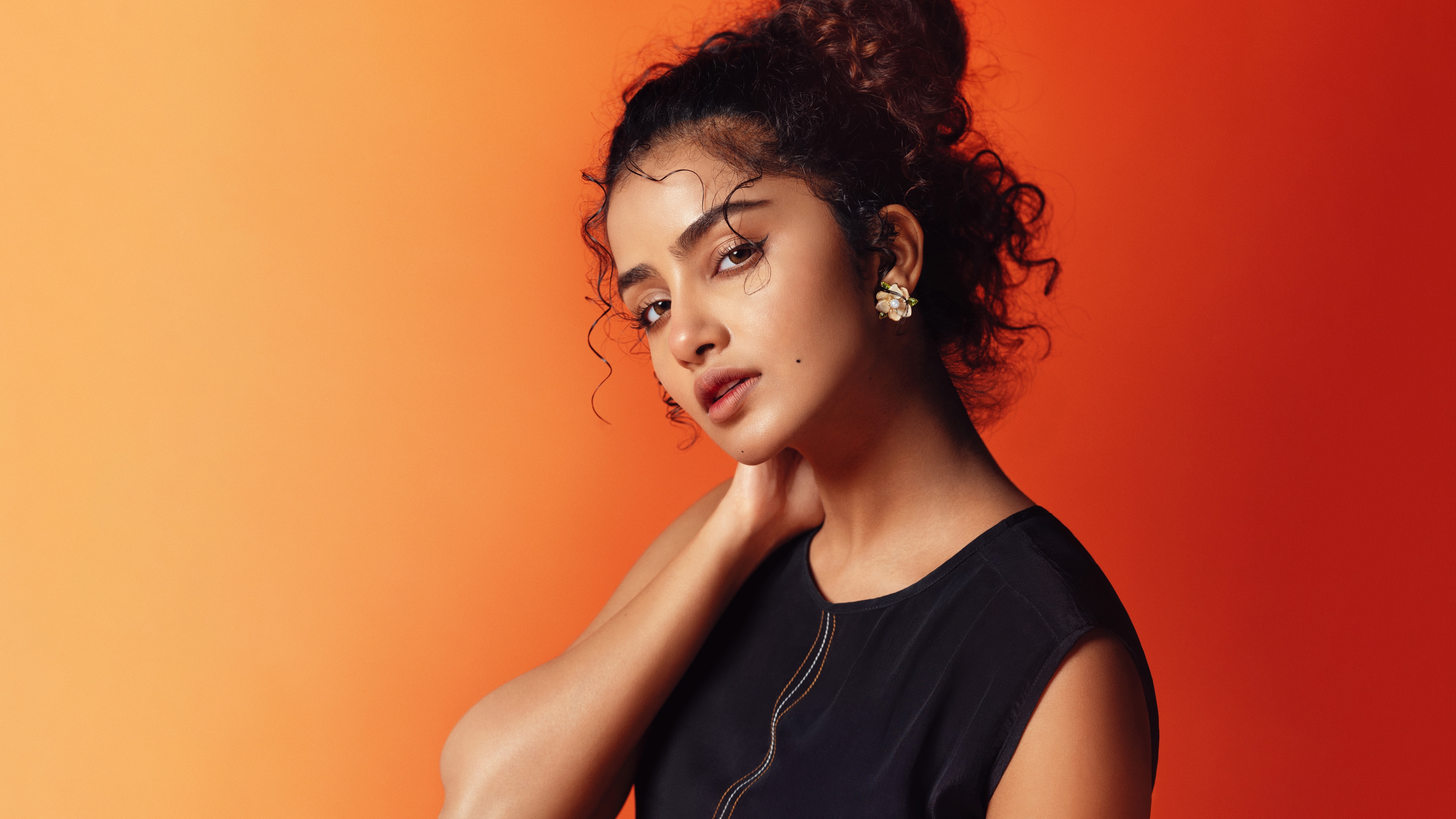 A high-definition portrait of Indian actress Anupama Parameswaran set against a bright orange background.