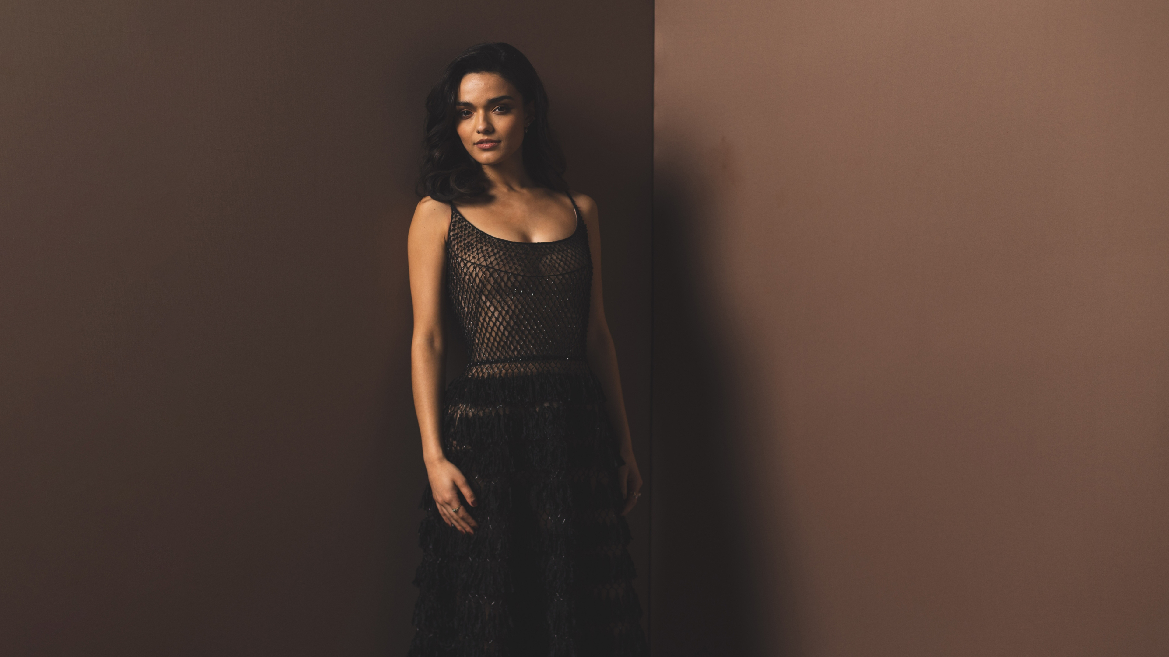 A professional close-up portrait of actress Rachel Zegler with a solid brown background.