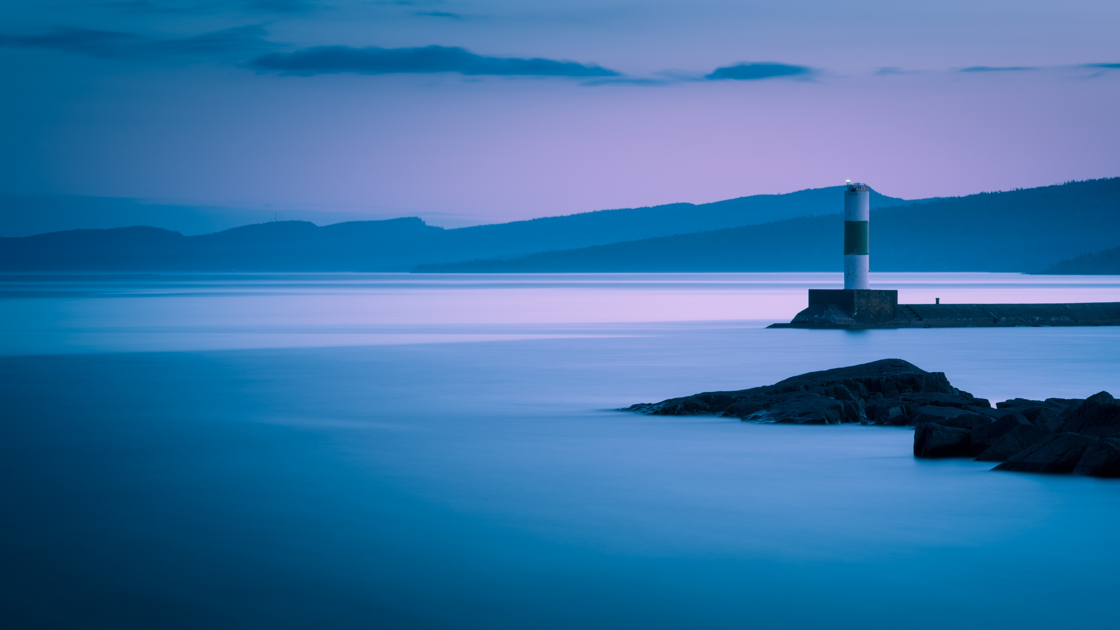 A long exposure photograph of a lighthouse on the Grand Marais coastline during dusk, featuring mountains and misty breakwaters.