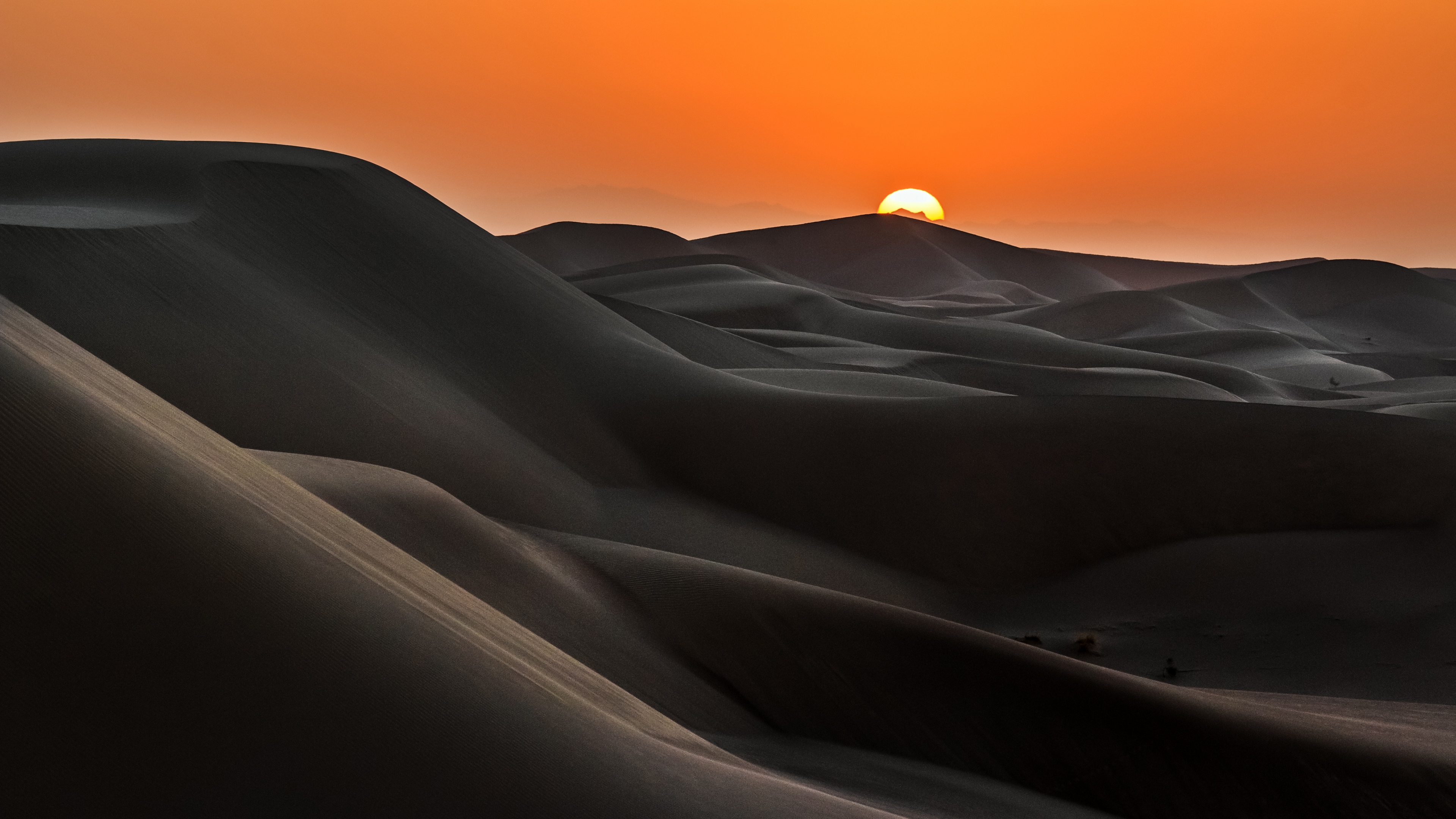 A serene desert landscape featuring rolling sand dunes under a bright orange sunrise sky during golden hour.