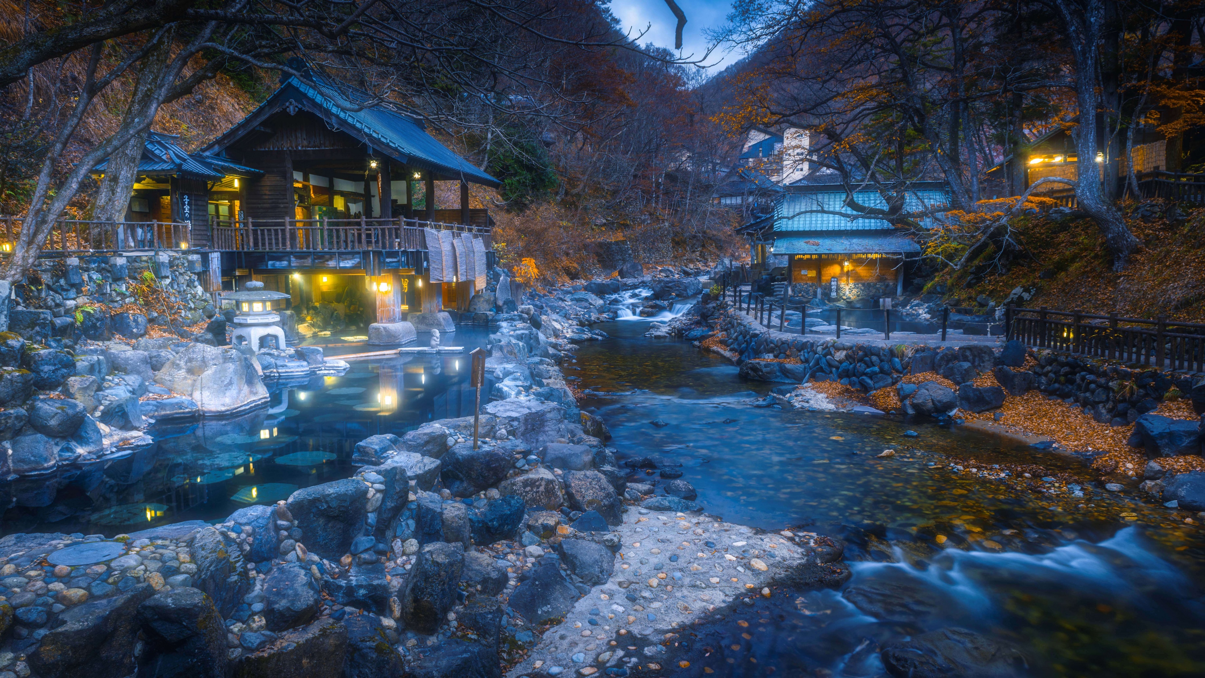An illuminated traditional Japanese outdoor hot spring surrounded by colorful autumn maple leaves at twilight.