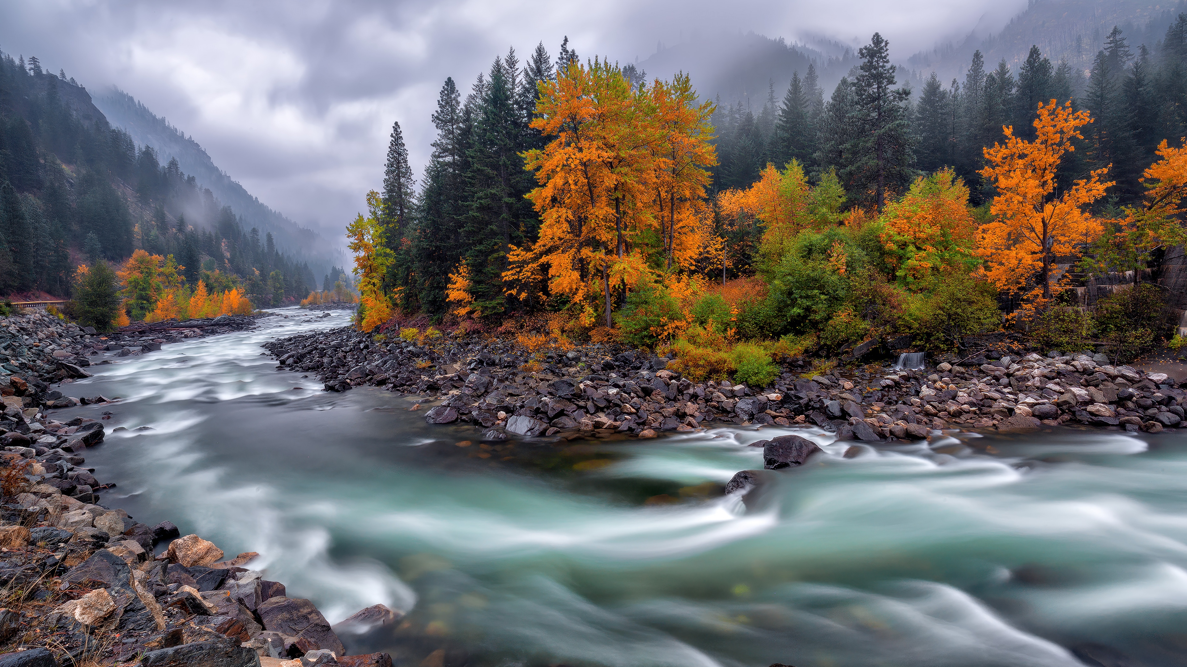 A long exposure photograph of a flowing river stream with autumn leaves and misty mountains in the background.