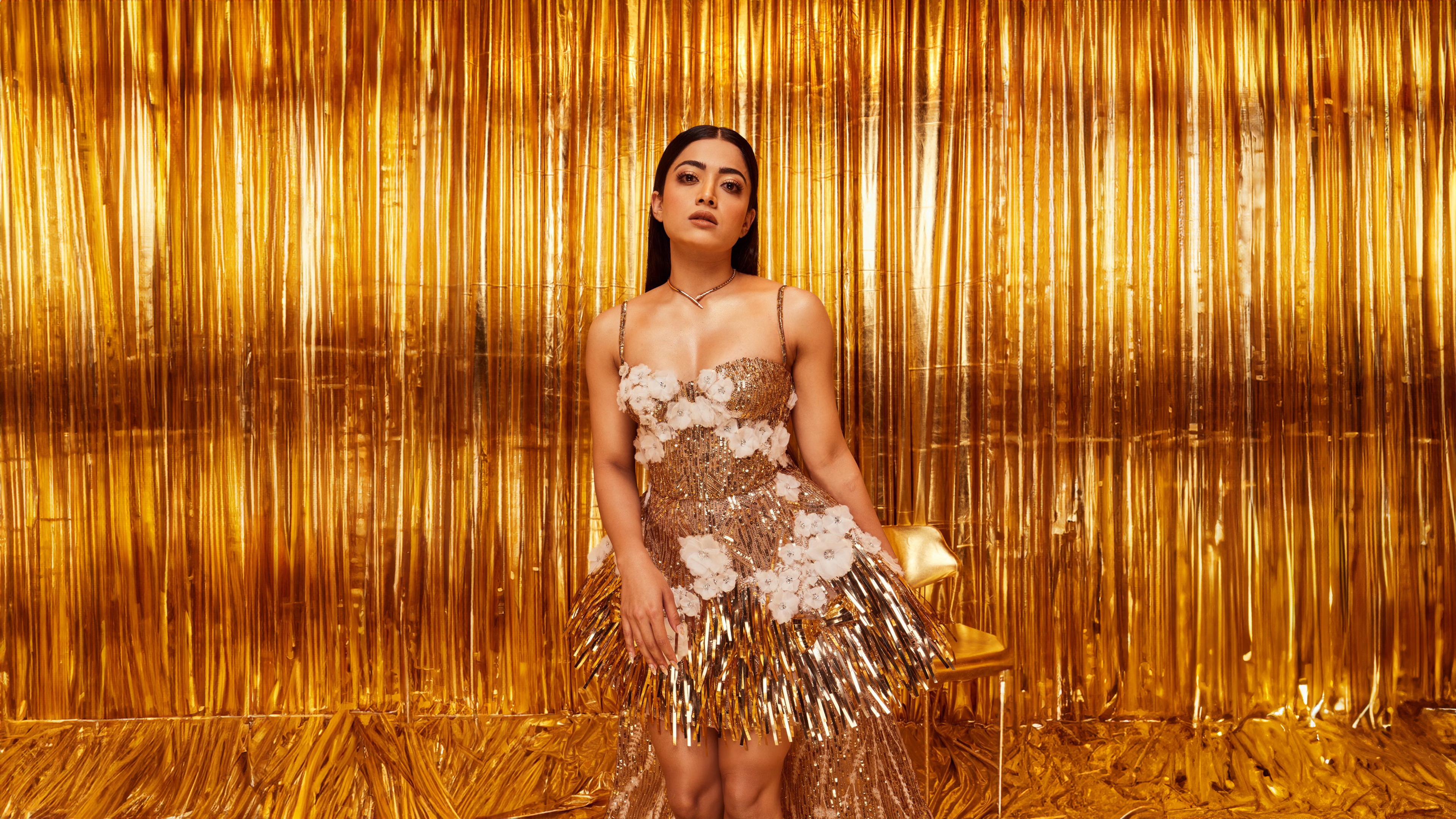 A high-glamour portrait of Indian actress Rashmika Mandanna against a radiant golden background.
