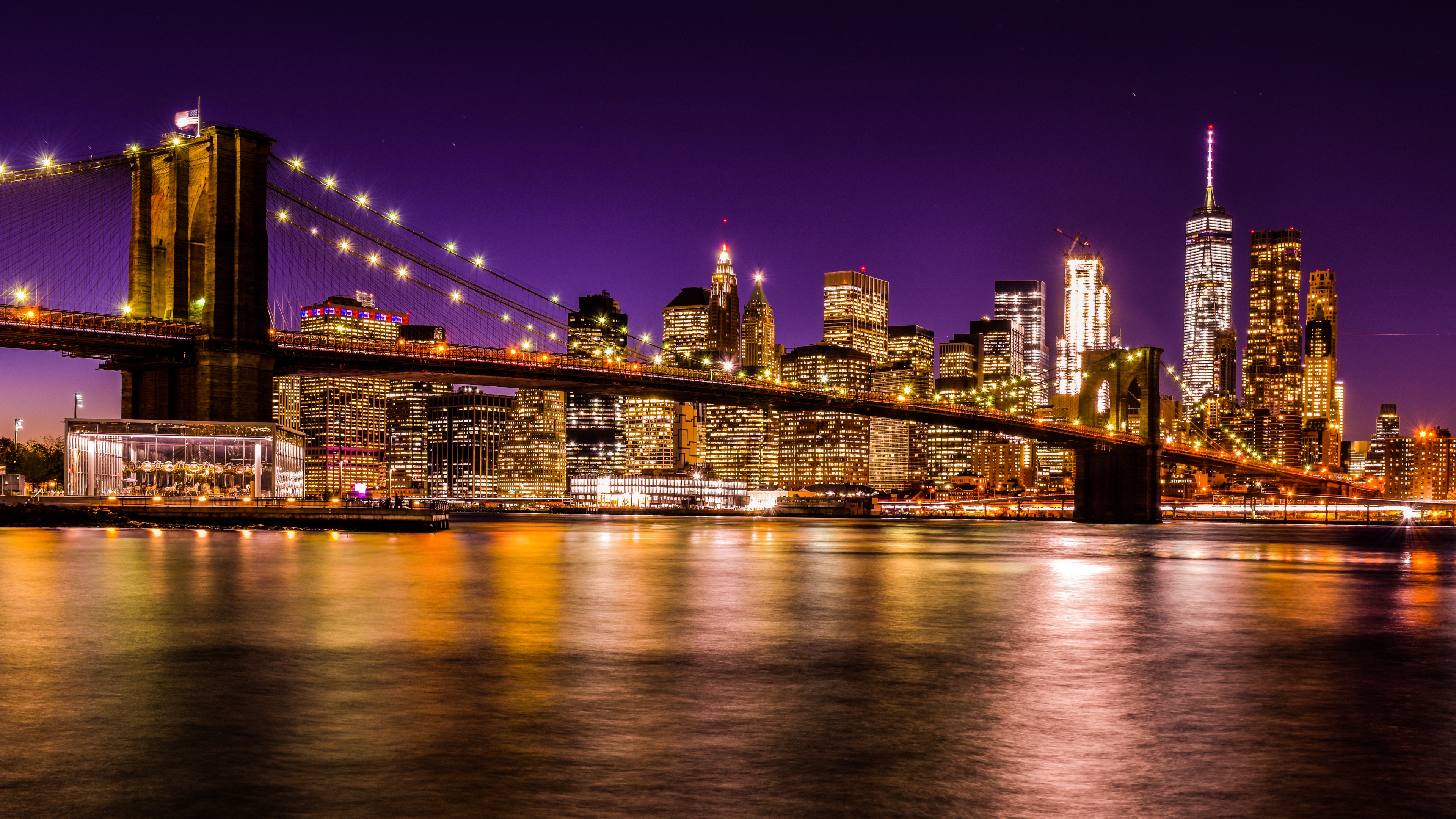 A long-exposure nighttime photograph of the Brooklyn Bridge and the New York City skyline under a purple sunset sky with reflections on the water.