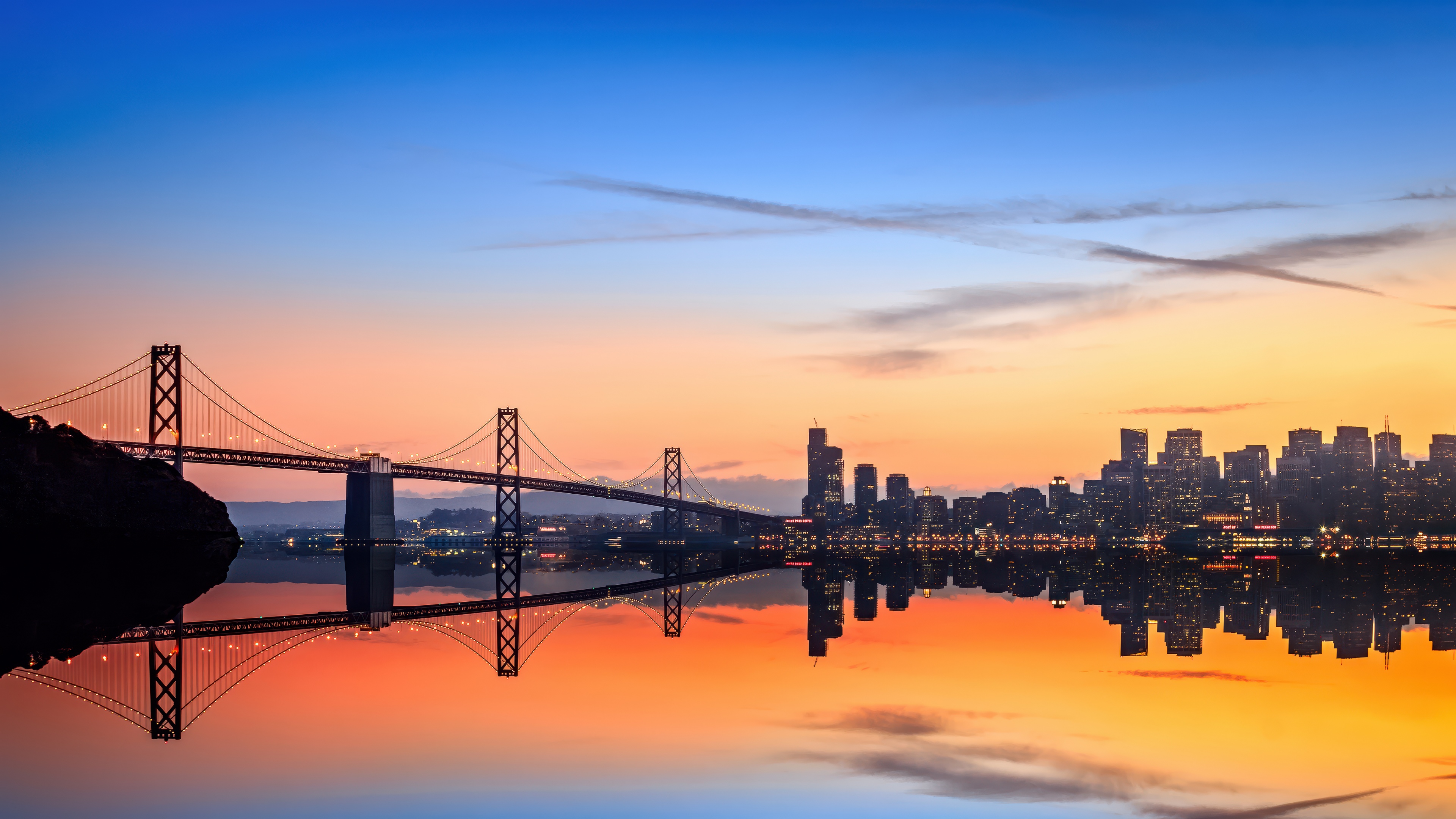 A panoramic view of the San Francisco-Oakland Bay Bridge at sunset with golden reflections on the water and a sprawling city skyline.